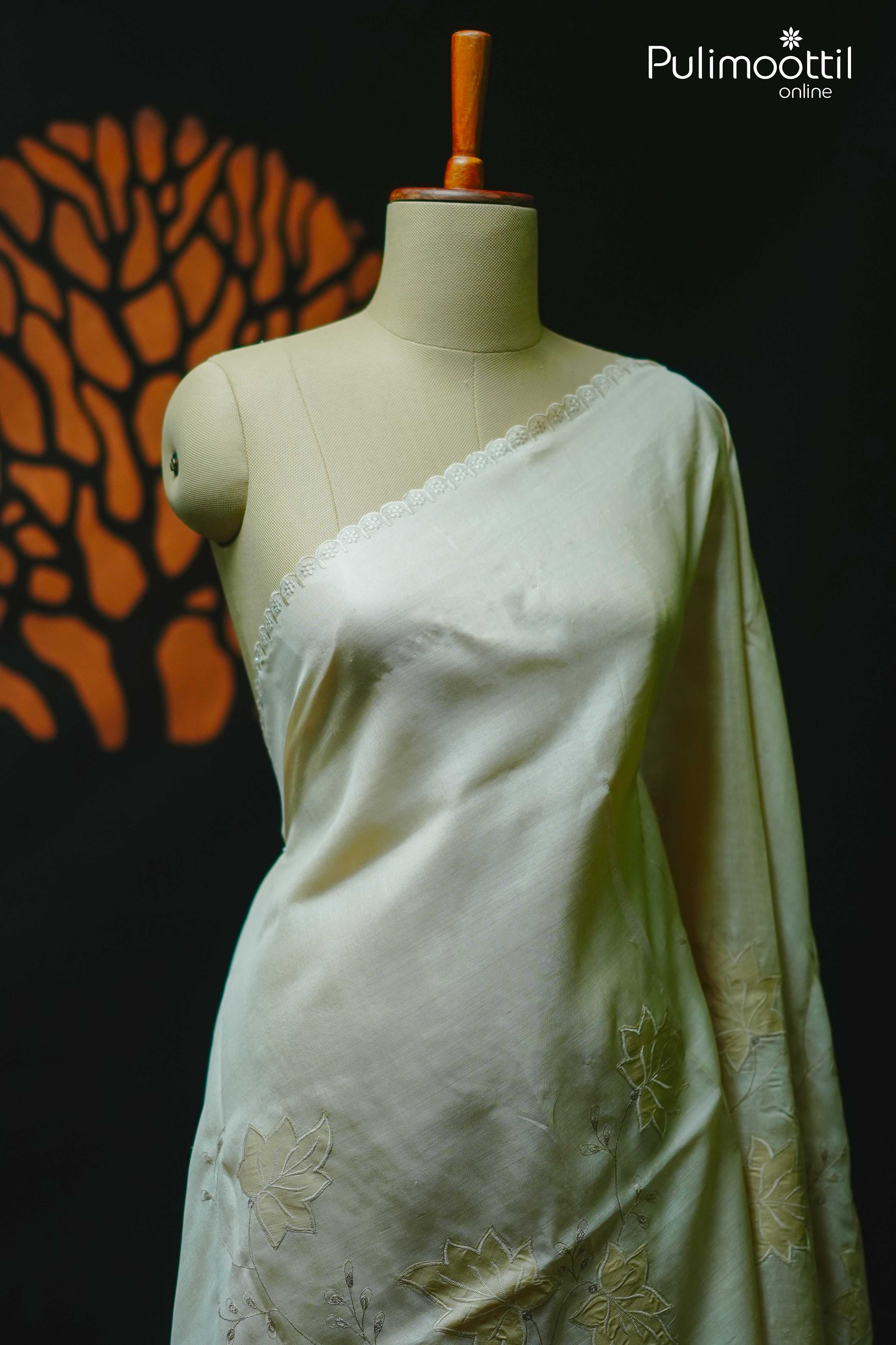 Off-white Tussar silk saree with delicate floral applique and scallop border, draped on a mannequin against a dark background with a tree design.