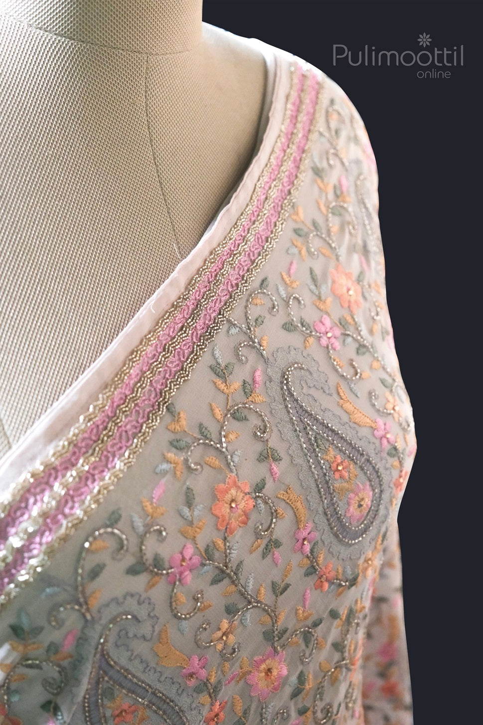 Close-up of pastel floral and paisley embroidery on light grey net saree with pink and gold embellished border displayed on a mannequin