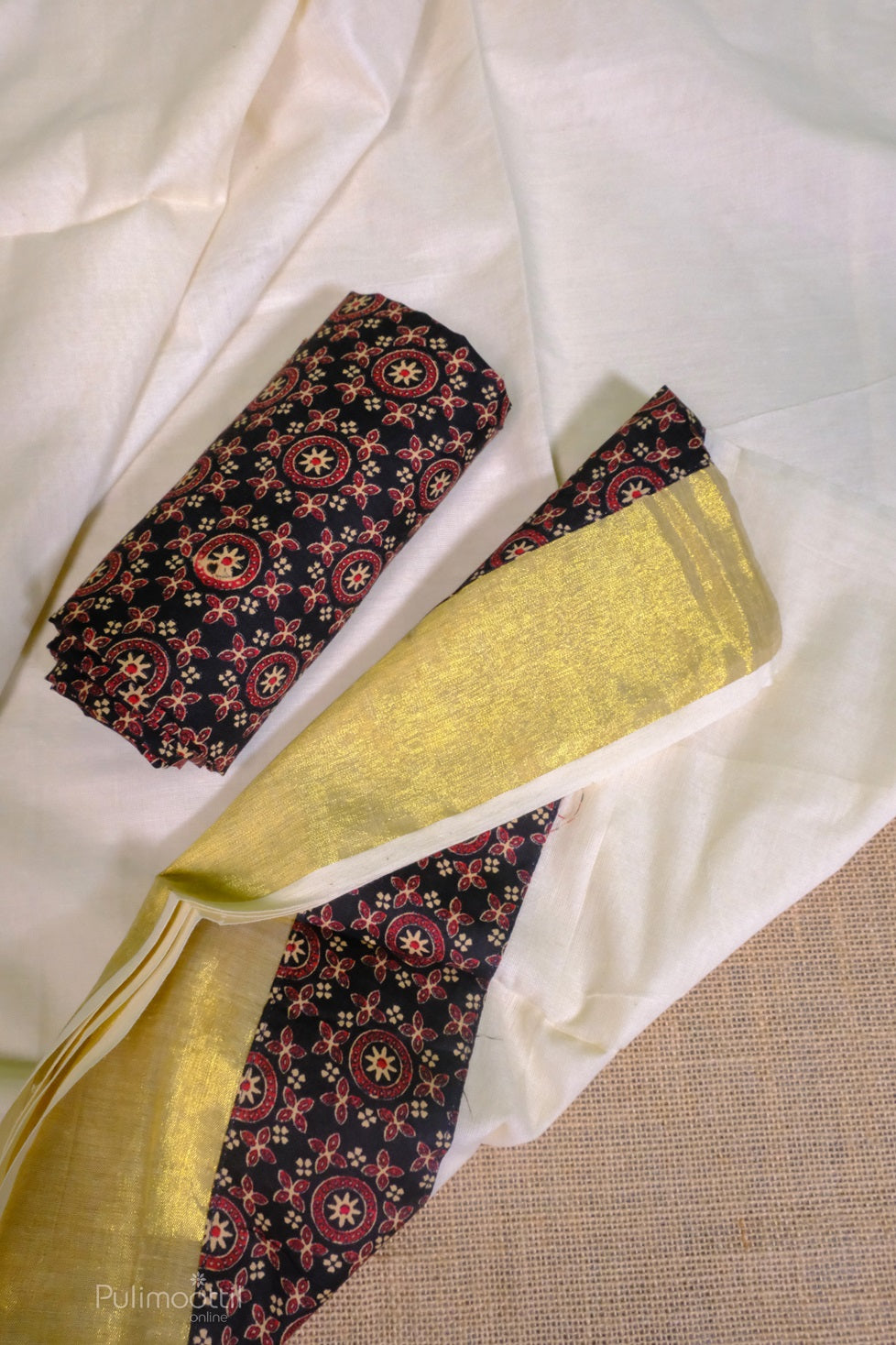 Rolled black printed blouse fabric placed beside the cream cotton saree with golden Kasavu border, laid on a jute surface.
