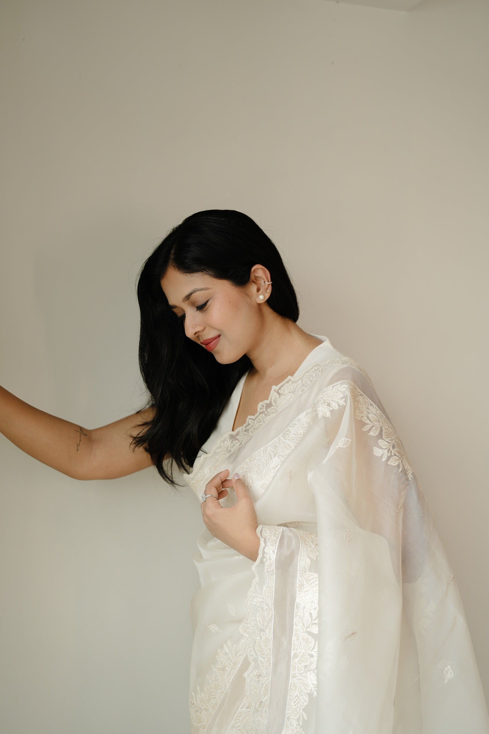 Woman in a white embroidered saree standing gracefully against a plain beige background, looking down with a soft smile and her hand gently holding the pallu.