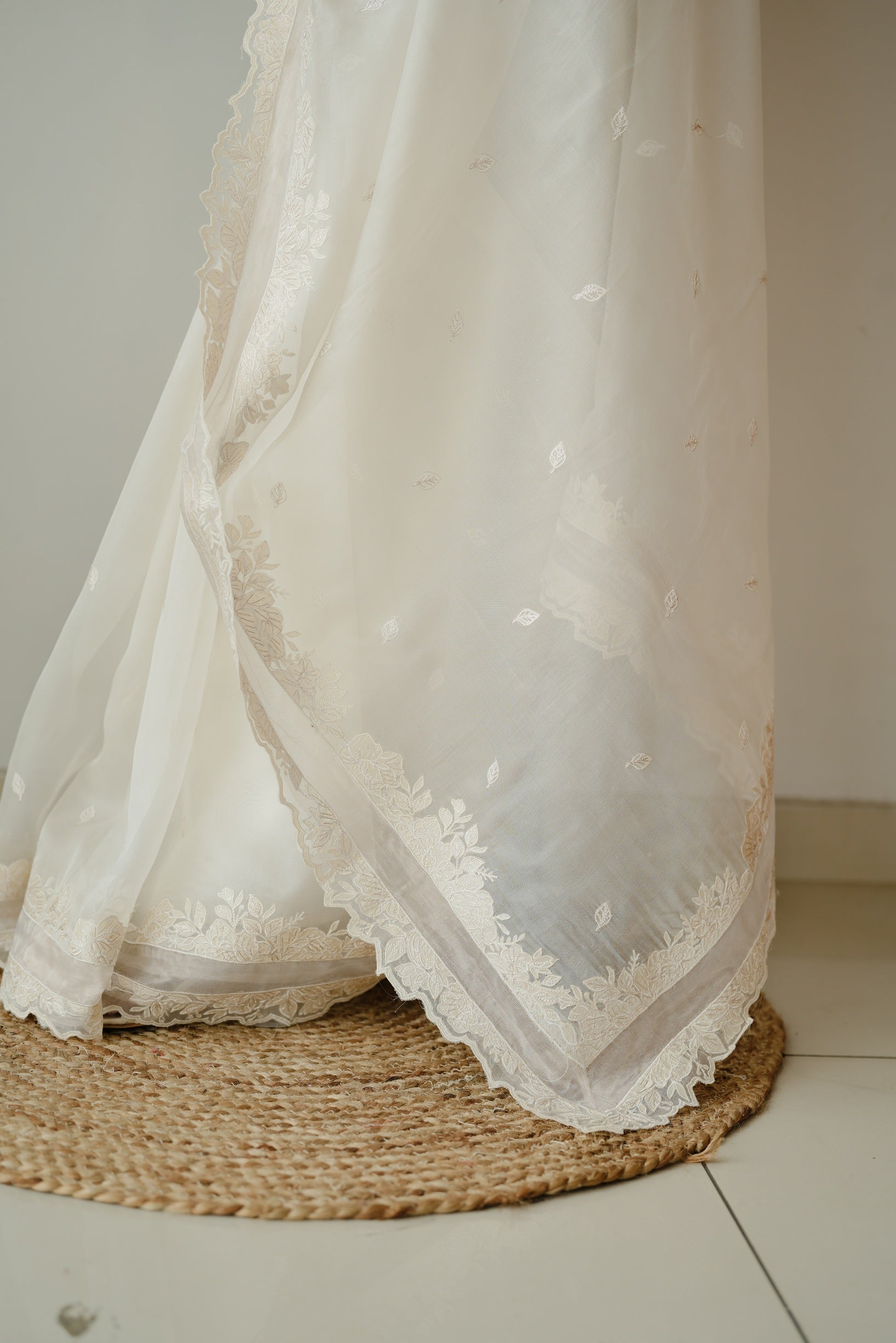 Close-up of the bottom part of a white saree with intricate floral embroidery along the border, displayed over a round woven mat.