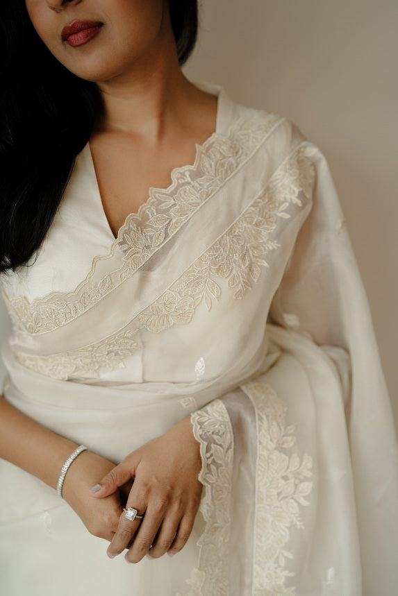 Mid-shot of a woman wearing a white saree, showcasing detailed floral embroidery near the neckline and pallu, with her hand resting near her waist adorned with a bracelet and ring.