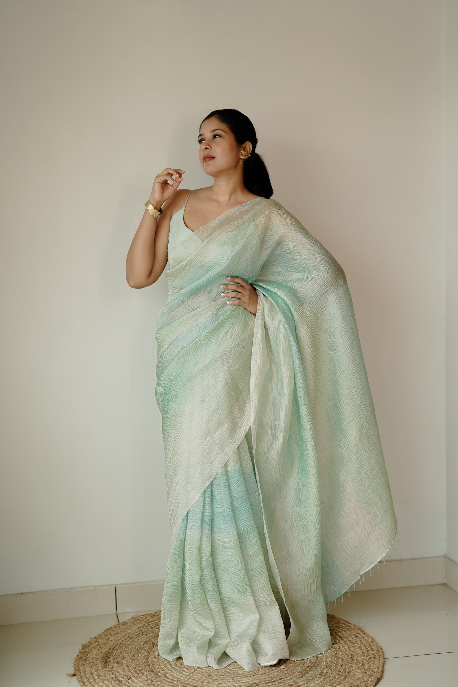 Full-length front view of a woman in a crinkled blue ombre Benarasi saree, standing barefoot on a round jute mat, exuding simplicity and elegance.