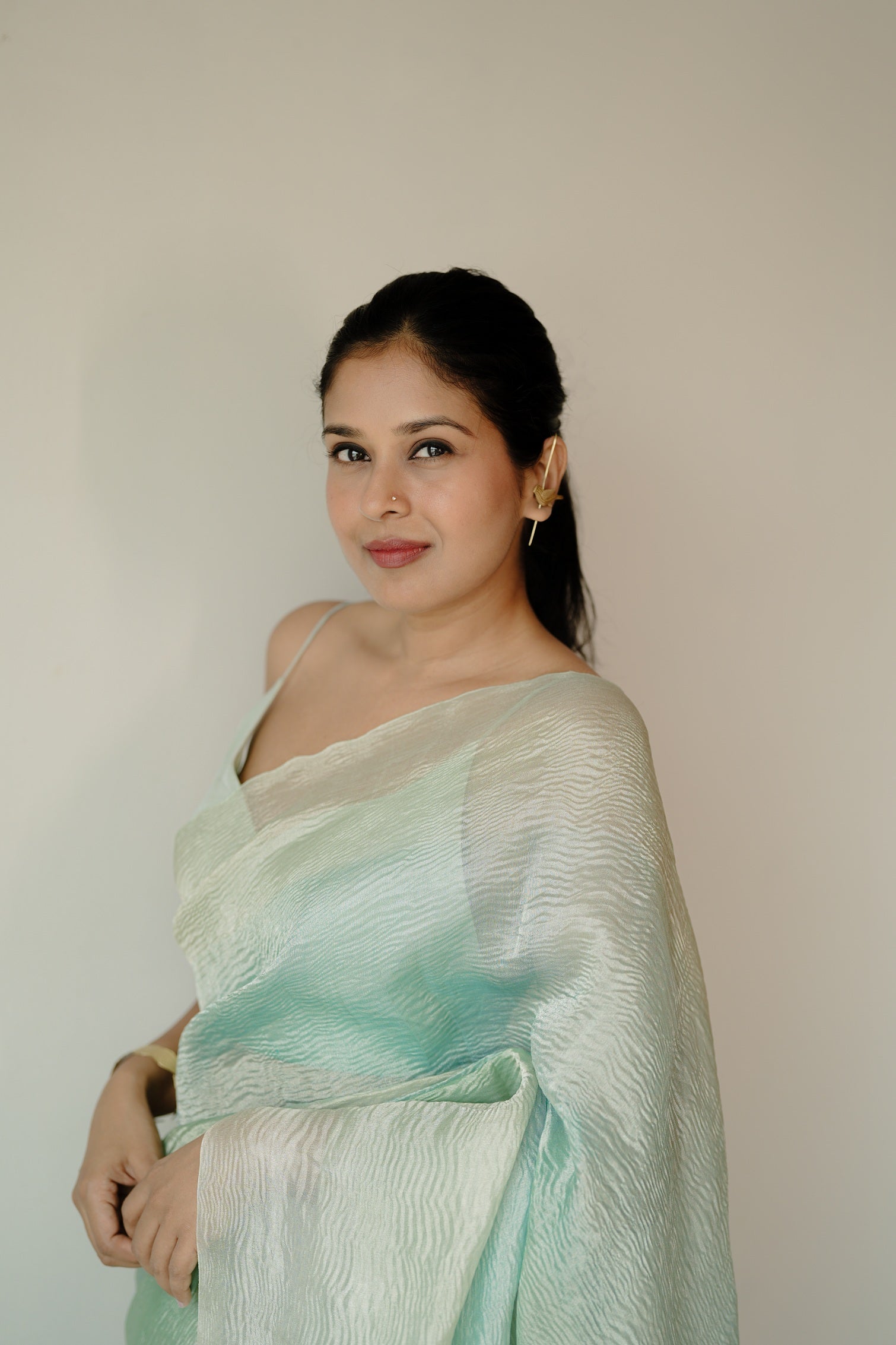 Side profile of a woman in a flowing blue ombre Benarasi saree, with soft pleats and a crinkled texture, looking over her shoulder in a serene indoor setting.