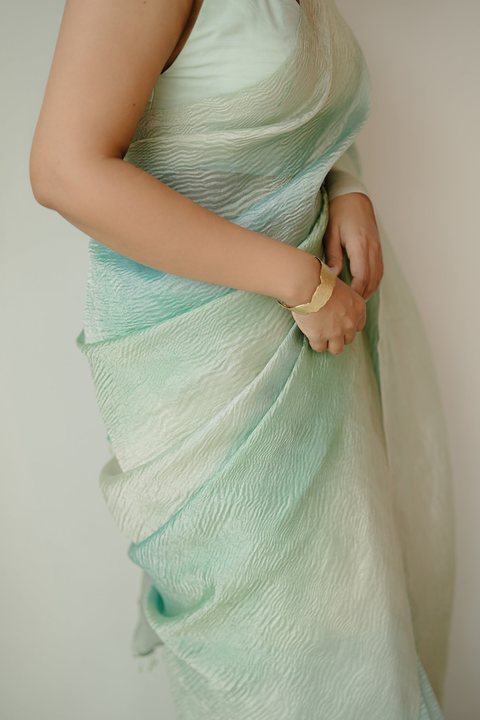 Rear view of a woman in a blue ombre crinkled Benarasi saree, showing the elegant drape of the pallu and her loose hairstyle, standing on a jute rug.