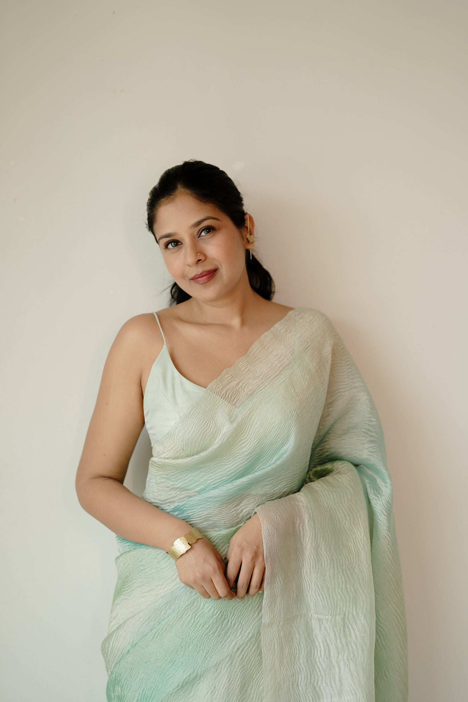 A woman stands gracefully in a blue ombre Pure Benarasi crush saree with crinkled texture, one hand raised to her head, posing against a minimalist cream background.