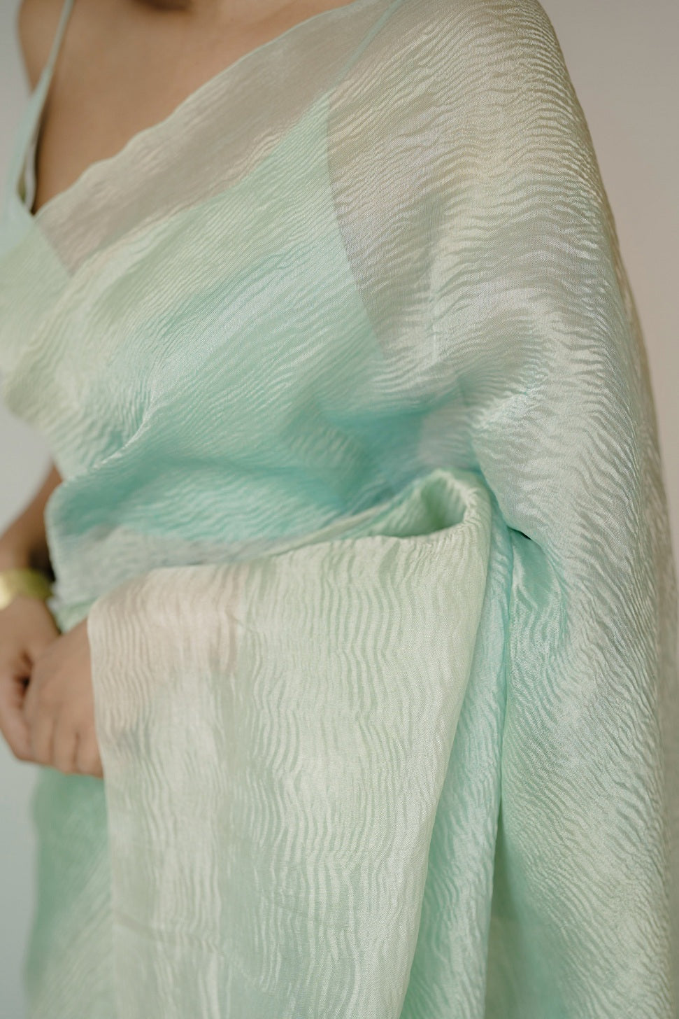 A woman elegantly adjusts her saree pleats while standing sideways, showcasing the soft crinkled texture and gradient of the blue ombre Benarasi fabric.