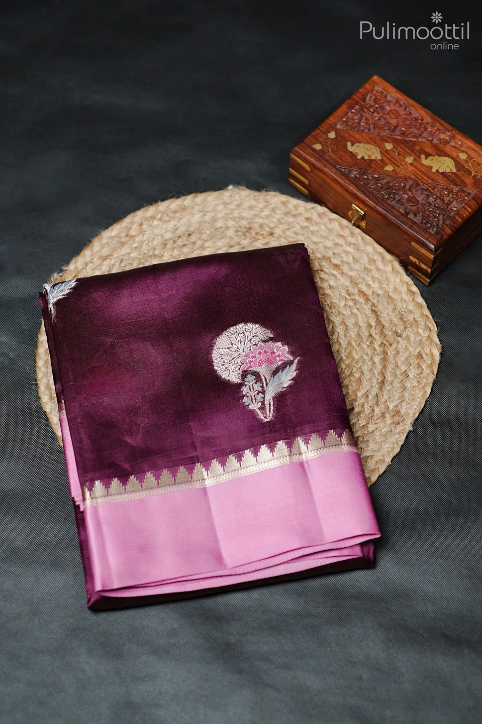 Purple mango silk saree with golden thread work and satin border, neatly folded on round thread mat with wooden box beside it.