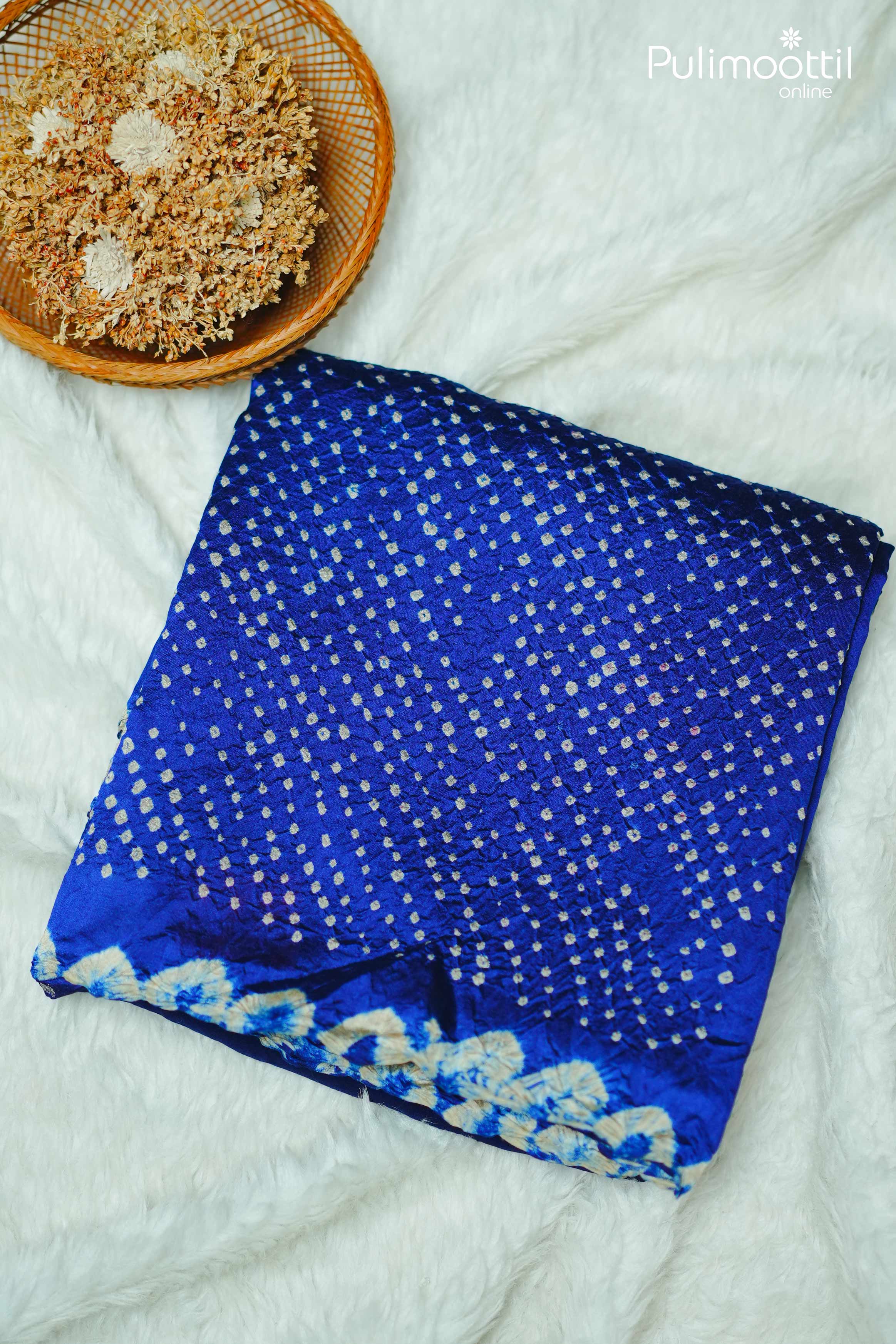 Folded royal blue Tussar silk saree with intricate white tie and dye designs and decorative border, placed next to a bowl of dried flowers.
