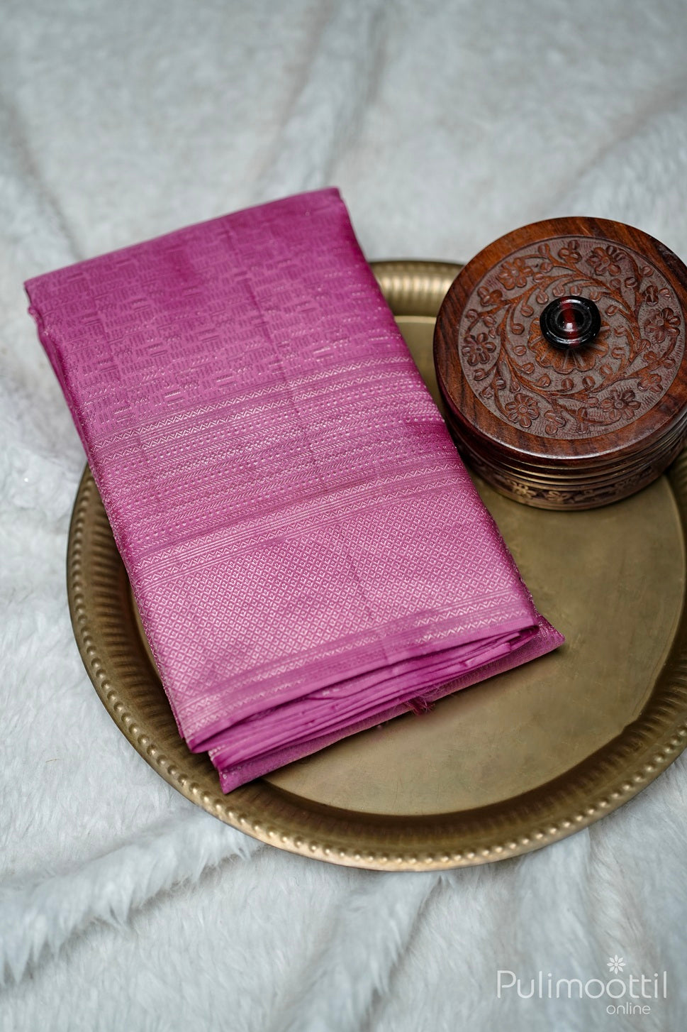 Soft pink silk saree folded on antique bronze plate with carved wooden box beside it.