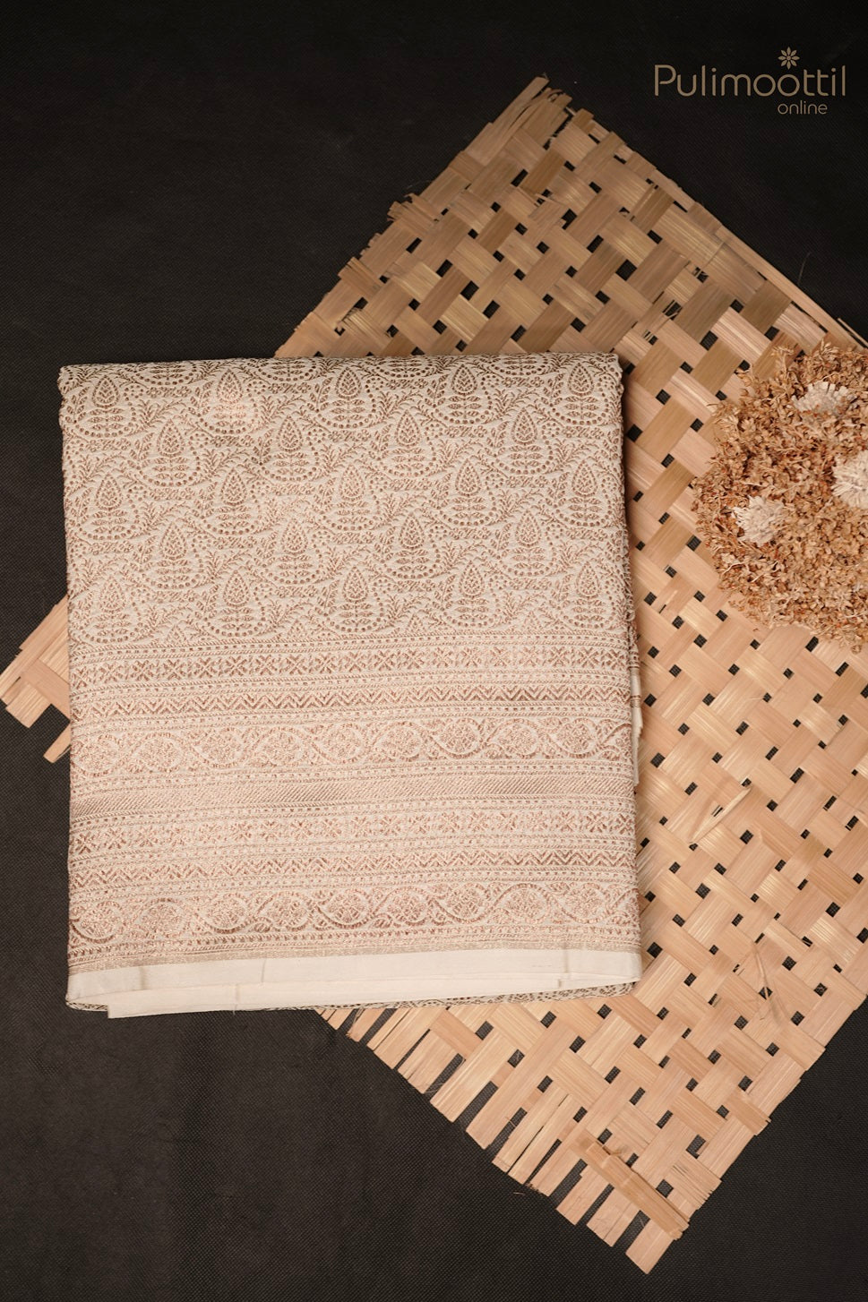Folded white Kattan silk saree with golden and thread floral motifs placed on woven mat with dried flowers