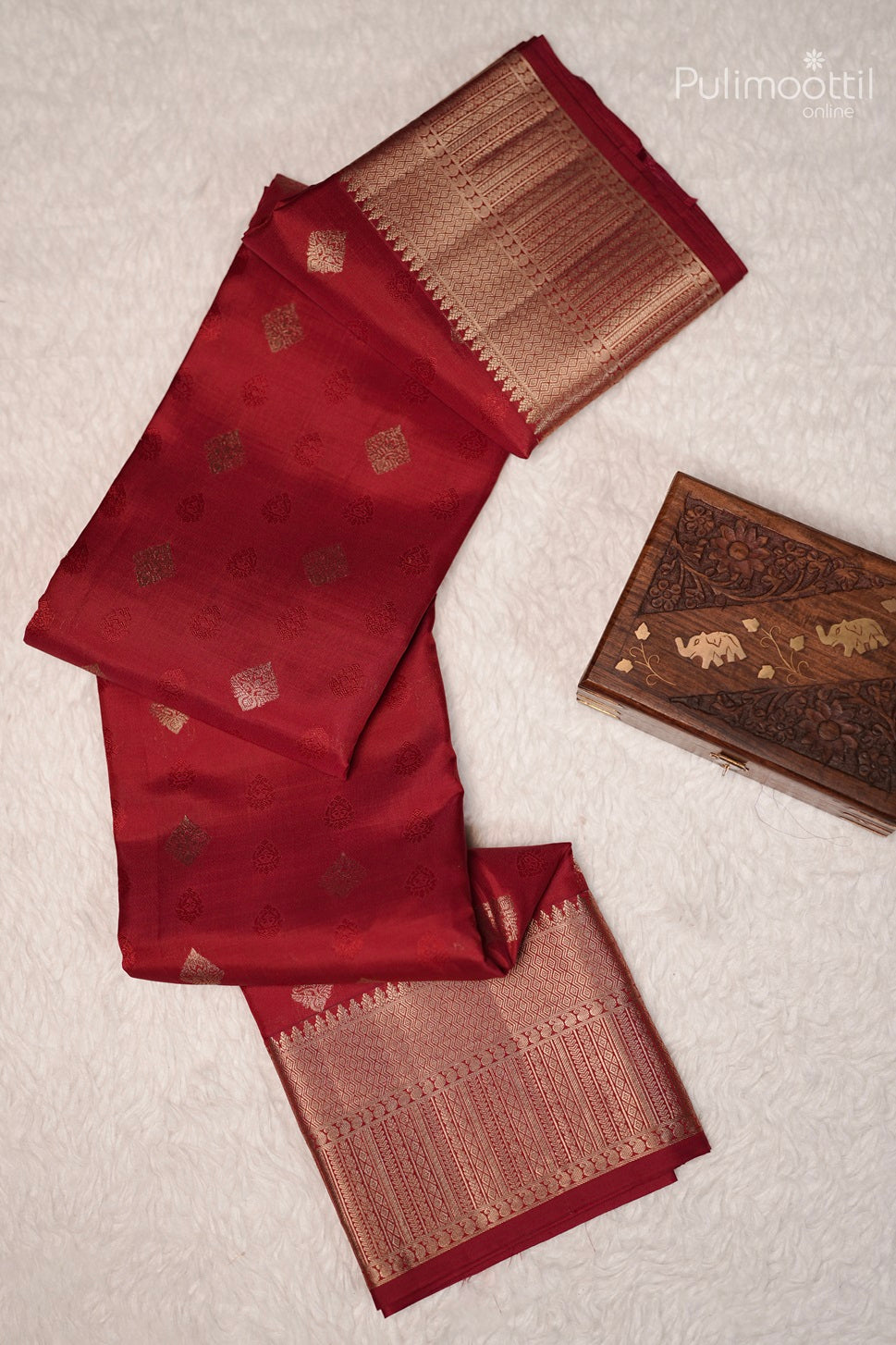 Folded chilli red Kanchipuram saree with golden buttas and border placed on a woven mat.