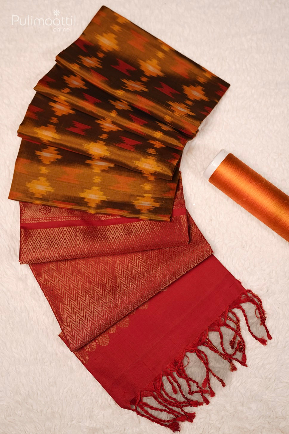 Ikat print fabric with red and orange hues, featuring a red zig-zag patterned border and tassels, along with a matching orange thread spool.
