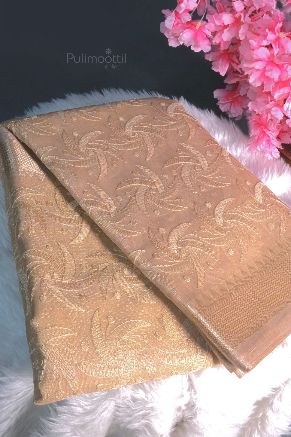 Chiku color Banaras tissue silk saree with intricate floral embroidery, placed on white fur with pink flowers in the background.