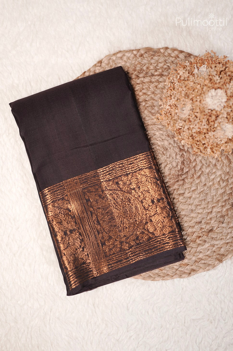 Folded black Kanchipuram soft silk saree with gold zari border, placed on a woven mat with a dried flower bouquet.
