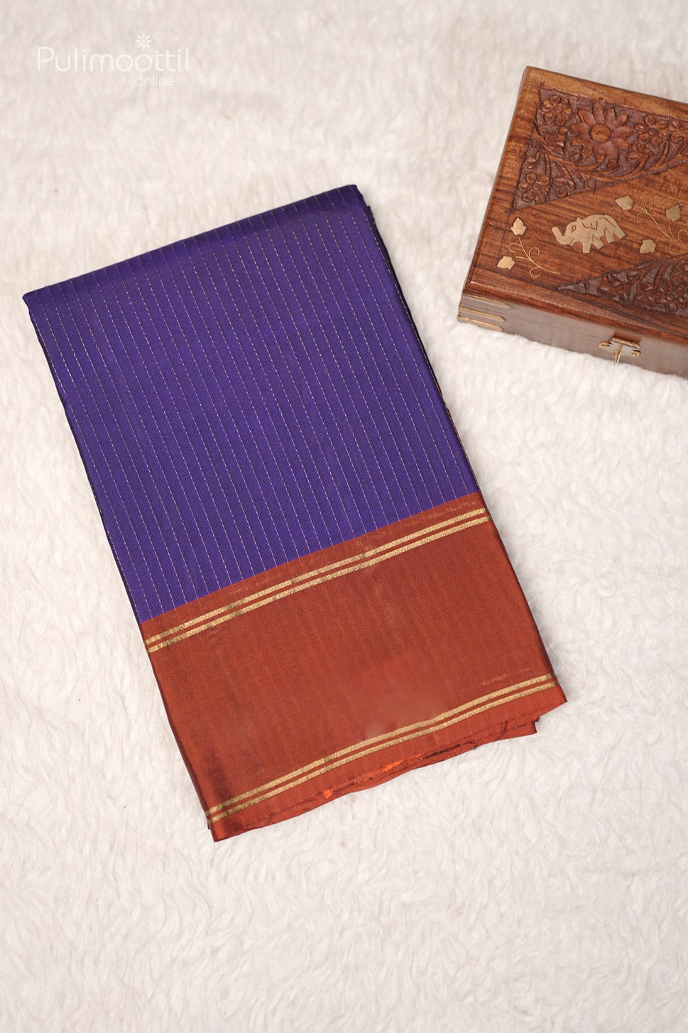 Folded violet saree with golden stripes and rust-colored border placed next to a carved wooden box on a white surface.