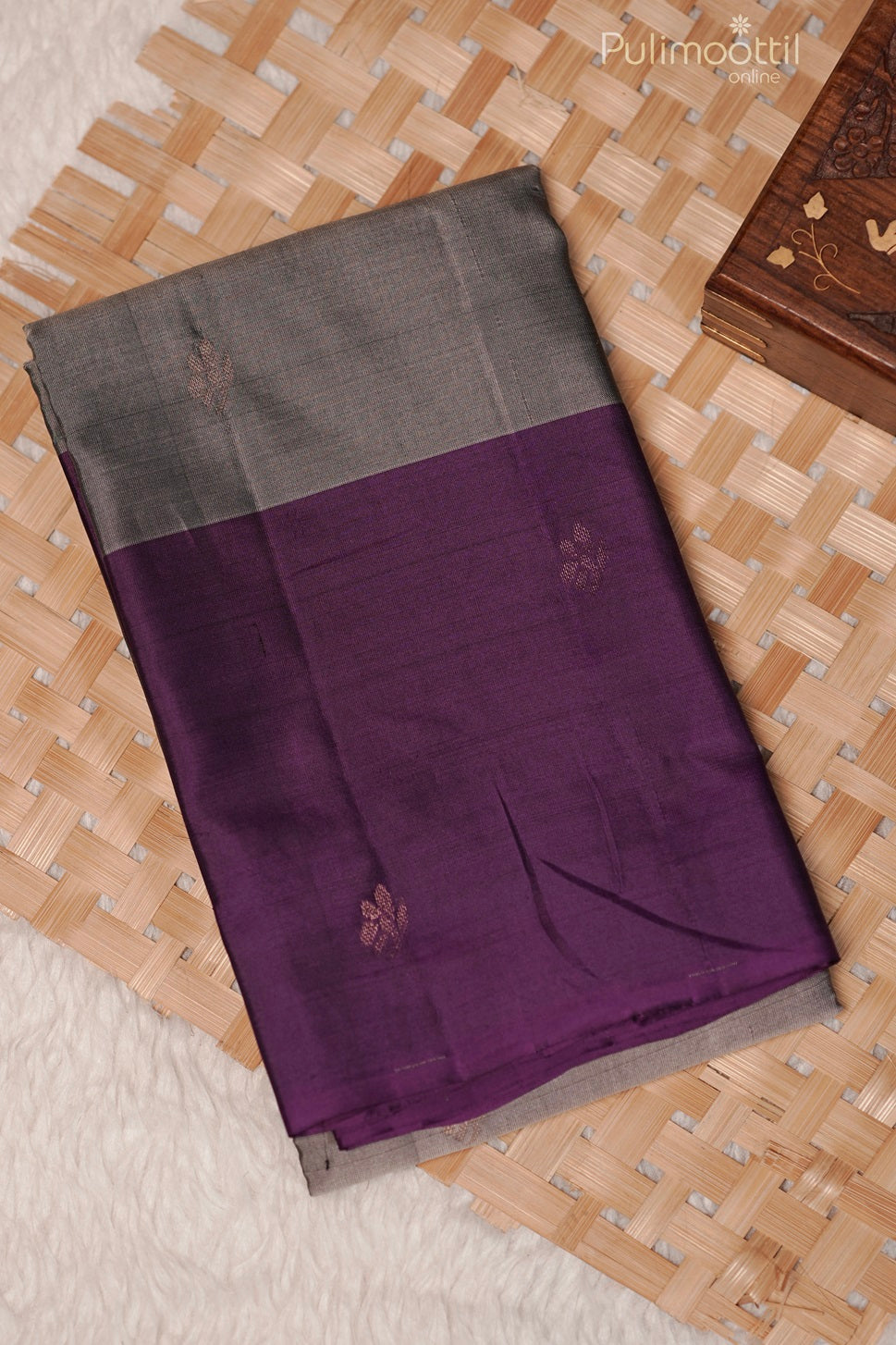 Folded grey Kanchipuram soft silk saree with purple contrast border and golden buttas, placed on a woven mat.