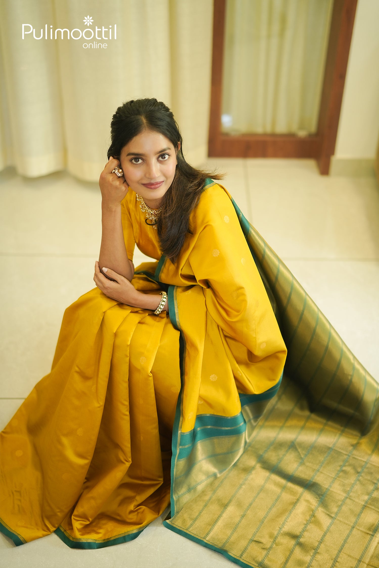 Lifestyle shot of the woman sitting, draping the Mustard Yellow Soft Silk Saree, showing the contrast Teal Green border