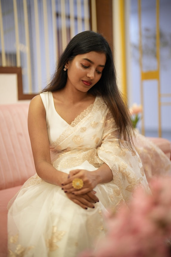 Woman posing serenely in a white organza saree with delicate floral embroidery and scalloped edges, seated near soft pink décor.