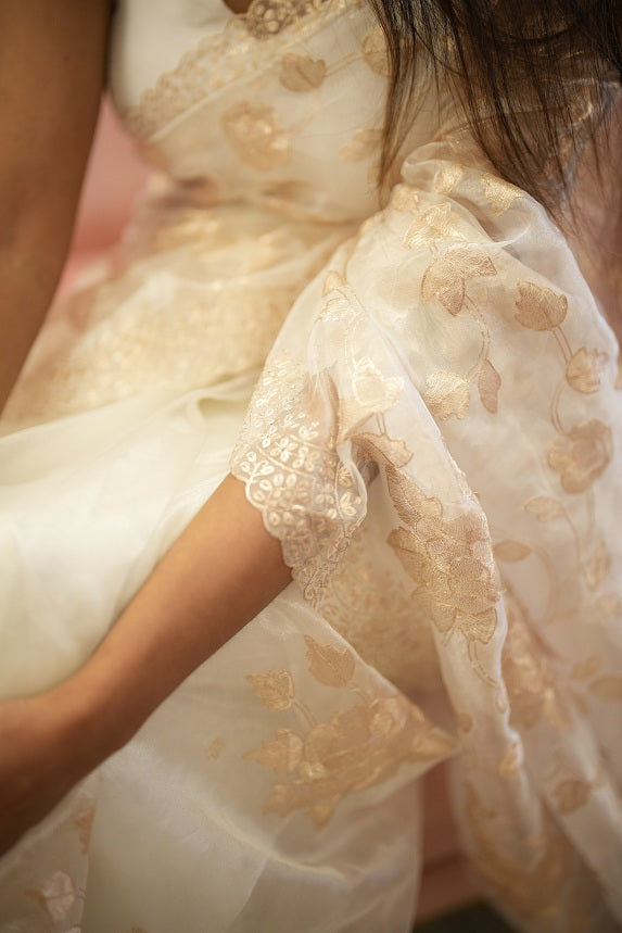 Elegant white organza saree with golden pitta floral embroidery and scallop border, draped gracefully by a woman