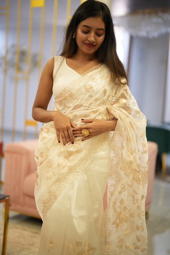 Model wearing a white organza saree with golden floral embroidery and scalloped lace border in a designer studio setting