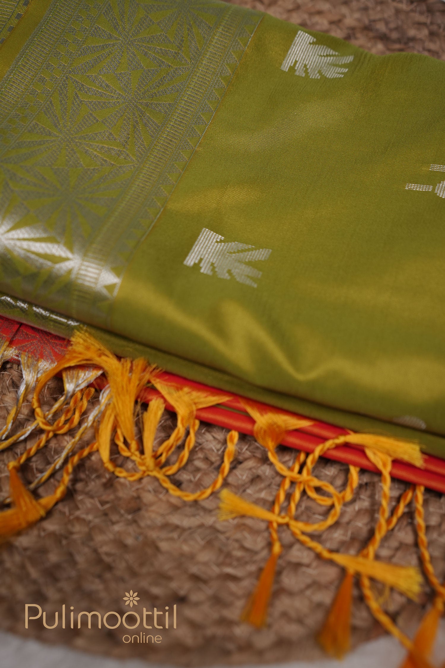Olive‑green semi‑silk saree with silver butta (small motif) detailing across the body and an intricately patterned silver border, softly folded with tassels at the edge, laid out on a woven mat.