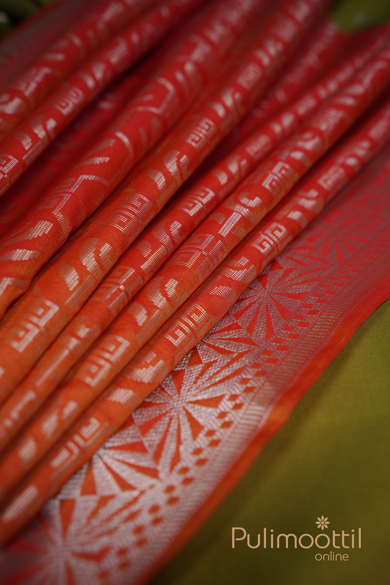 Olive green semi‑silk saree folded to showcase orange tassels, featuring silver butta motifs across the body and an intricate silver geometric border, styled on a woven mat.
