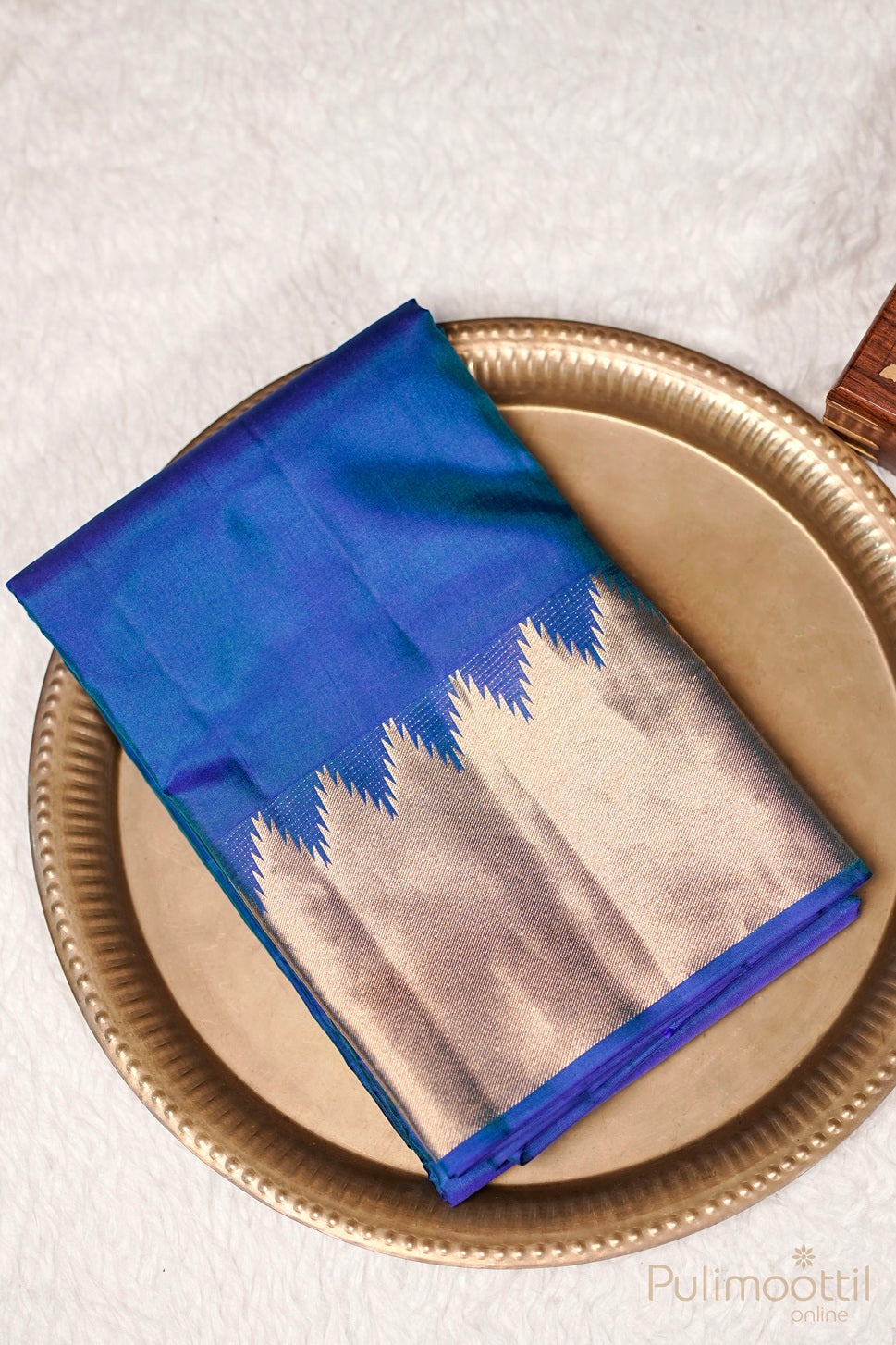 Peacock blue Kanchipuram saree with golden temple border neatly folded on a brass plate.