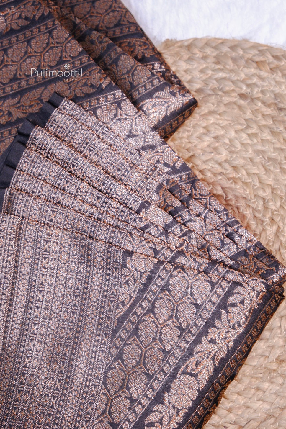 Close-up view of an intricately woven black and copper-brown saree with rich floral and geometric patterns, folded neatly over a textured beige mat.