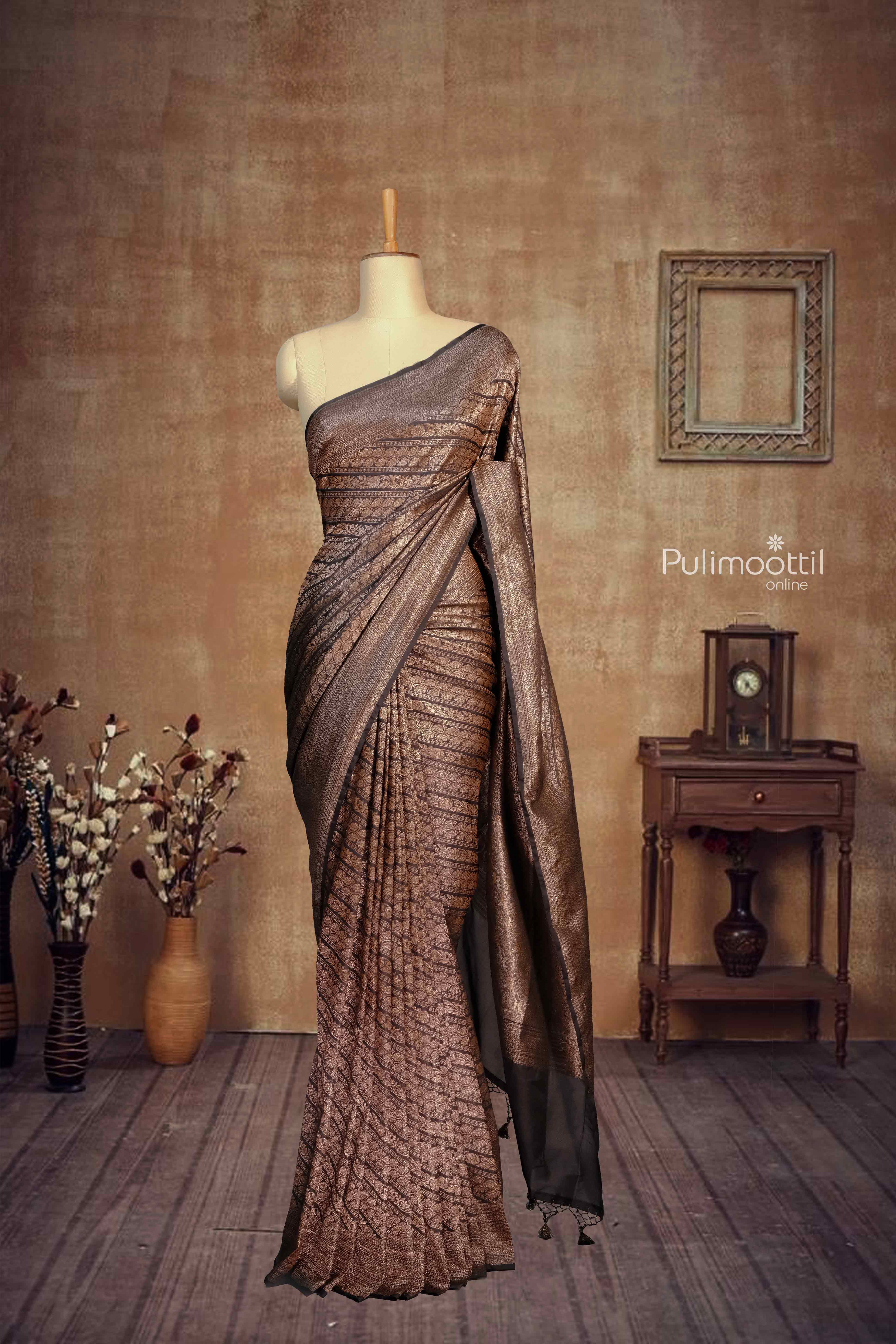 Elegant black and copper-brown traditional saree draped on a mannequin in a vintage-themed room with wooden flooring, vases with dried flowers, and a classic wall clock.