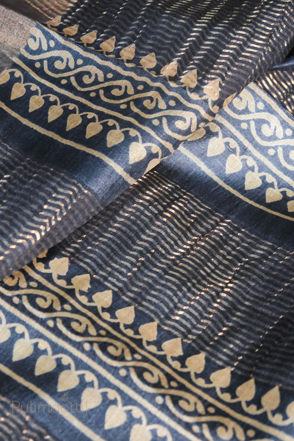 Close-up of black cotton silk fabric featuring intricate golden zari stripes and ornamental border with leaf and vine motifs, showcasing traditional craftsmanship.