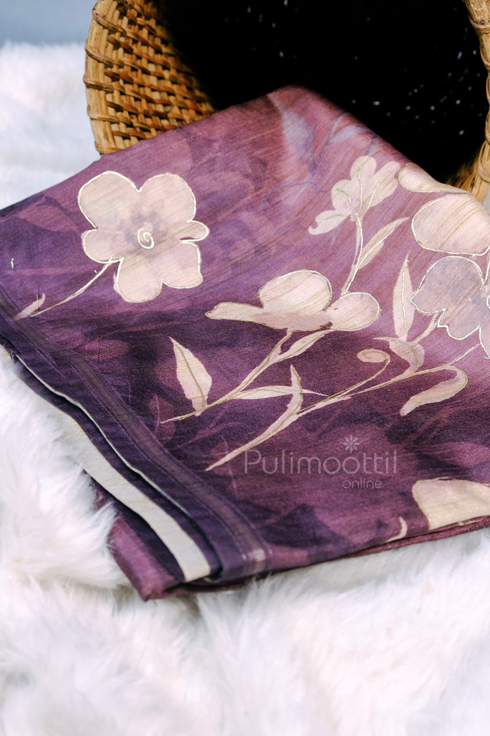 Folded purple silk designer saree with delicate cream floral prints, placed in a wicker basket on white faux fur.
