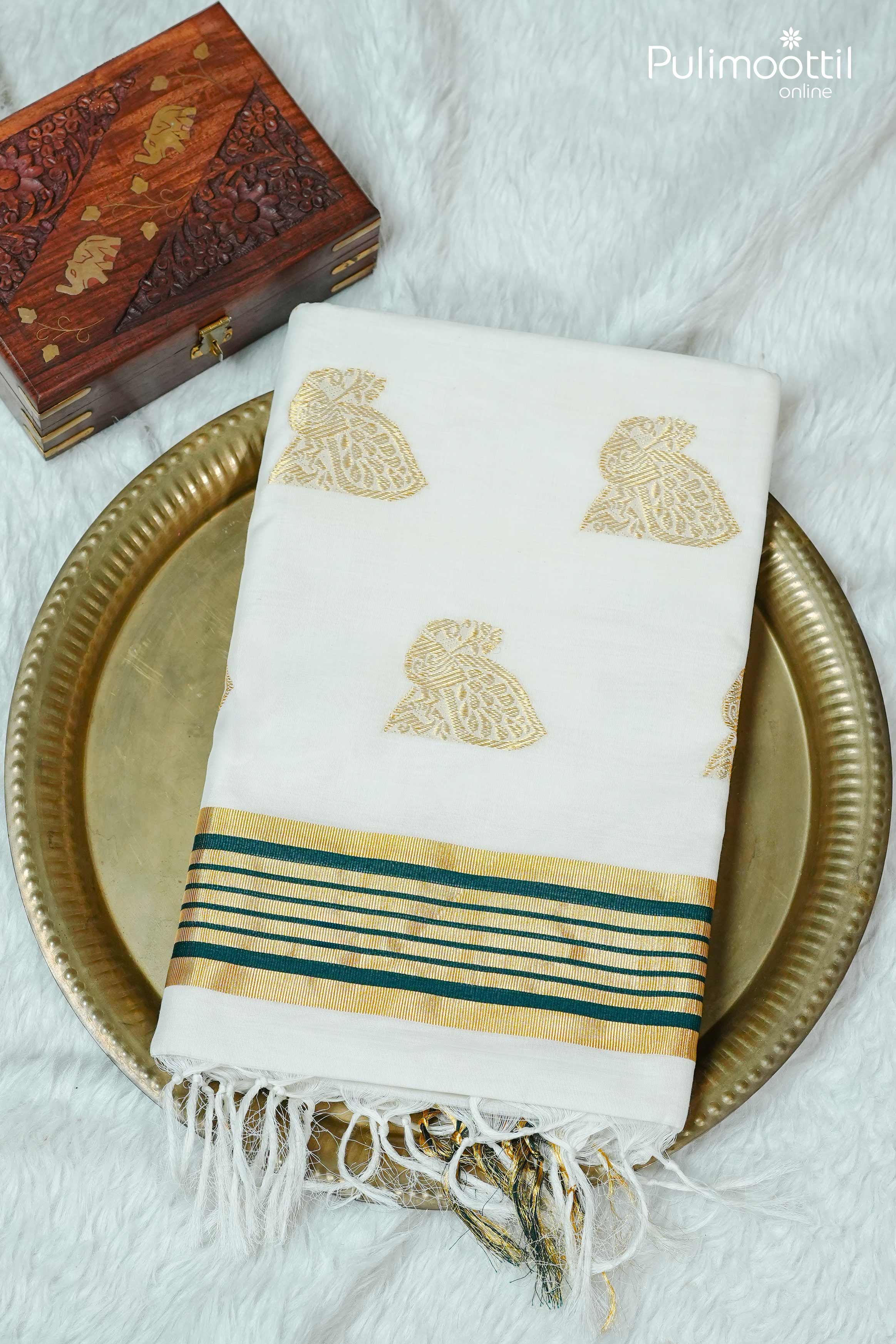  Folded cream colour Kerala set saree with golden zari peacock motifs and golden-green striped tissue border, placed on brass plate with wooden carved box for festive display.

