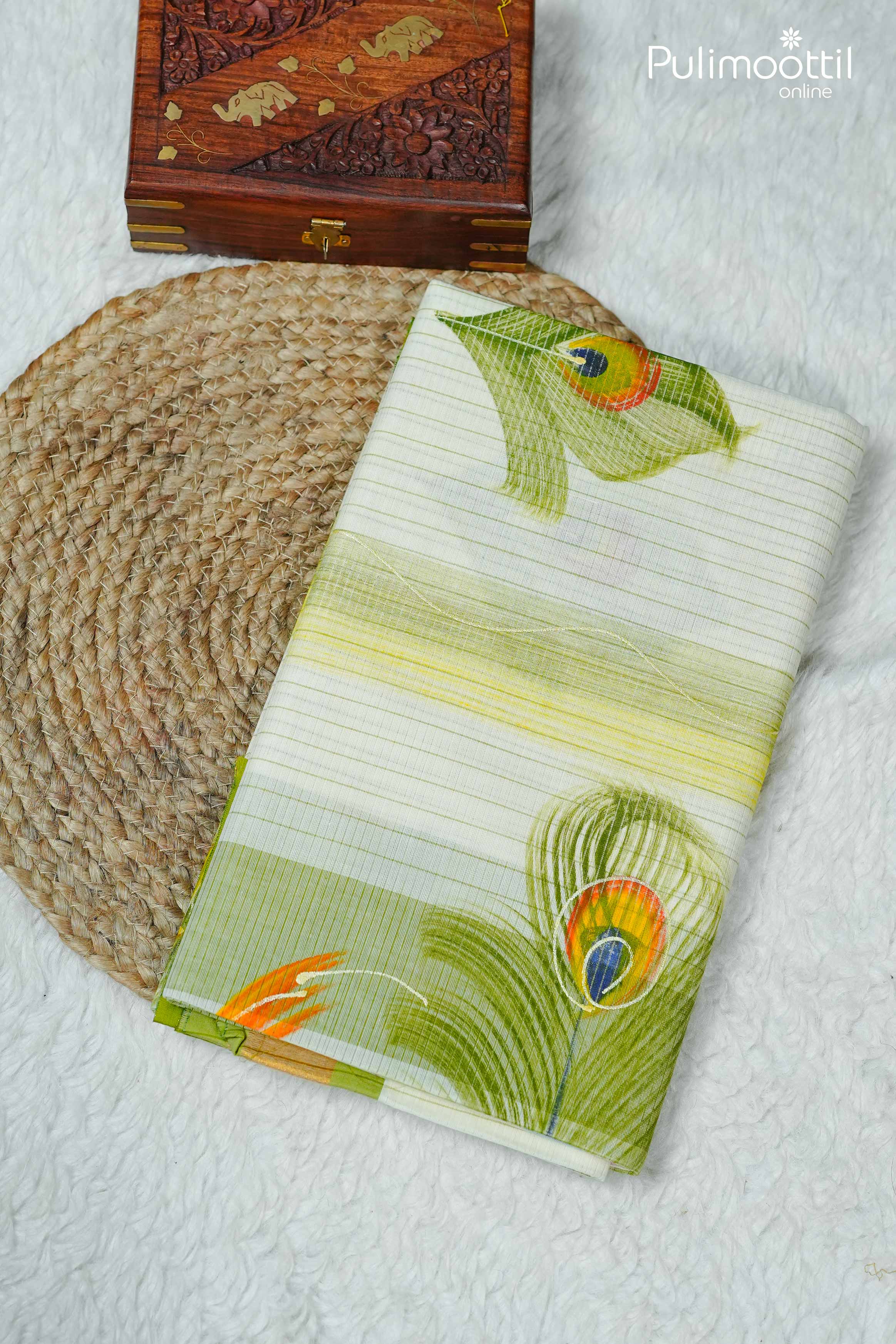 Folded cream Kerala traditional set saree with golden tissue border and hand-painted peacock feather motifs, placed on woven mat beside wooden jewelry box.