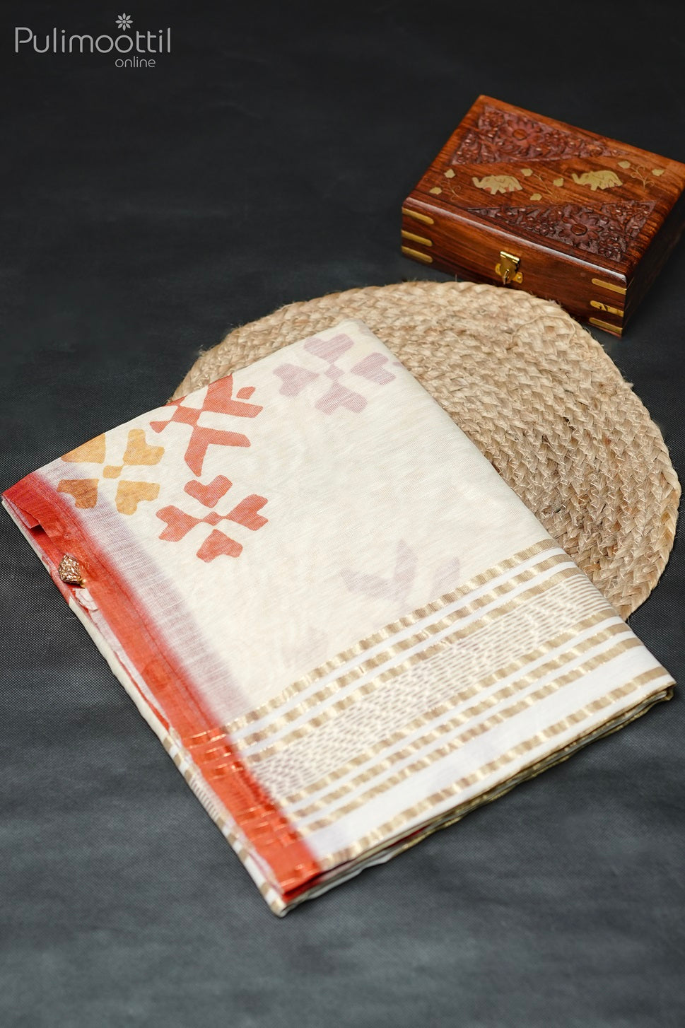 Cream net cotta saree with multicolor motifs, neatly folded on a wooden mat with a wooden box placed beside it.
