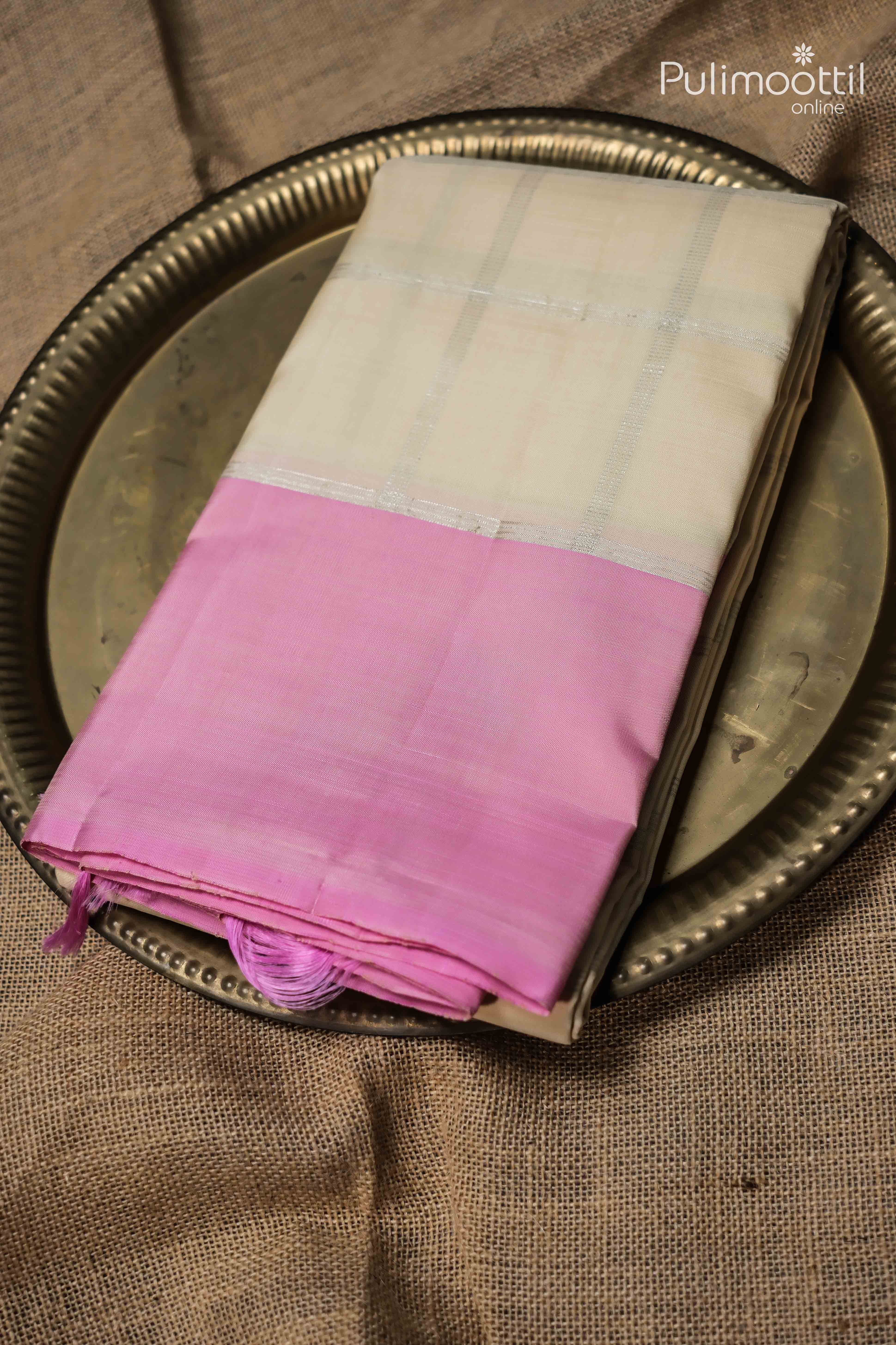 Folded cream colour pure Kanchipuram silk saree with silver checked design and soft pink border placed on a decorative brass plate.