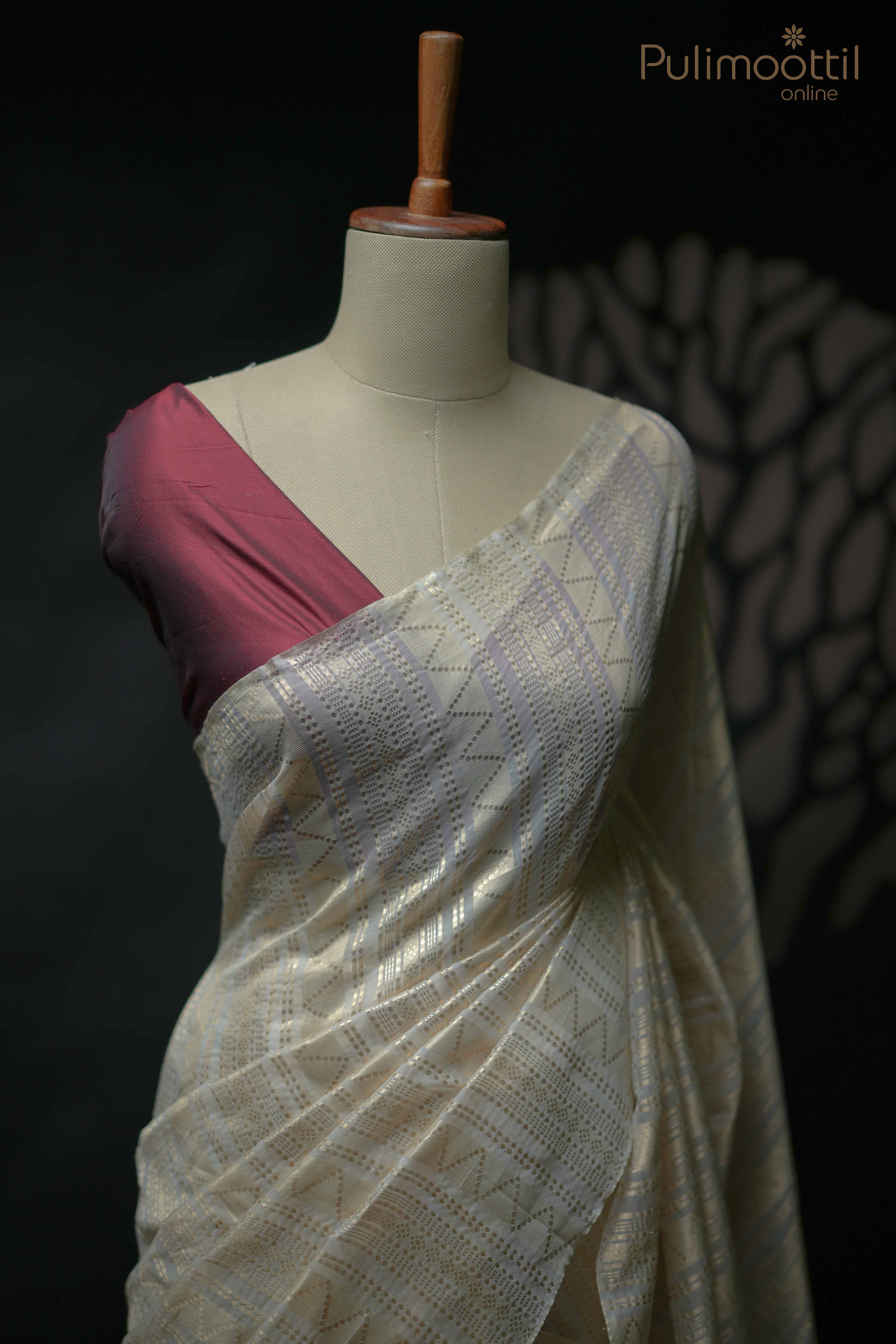 Cream-colored soft organza saree with golden zari design, draped on a mannequin and paired with contrasting maroon blouse fabric — elegant and festive look.