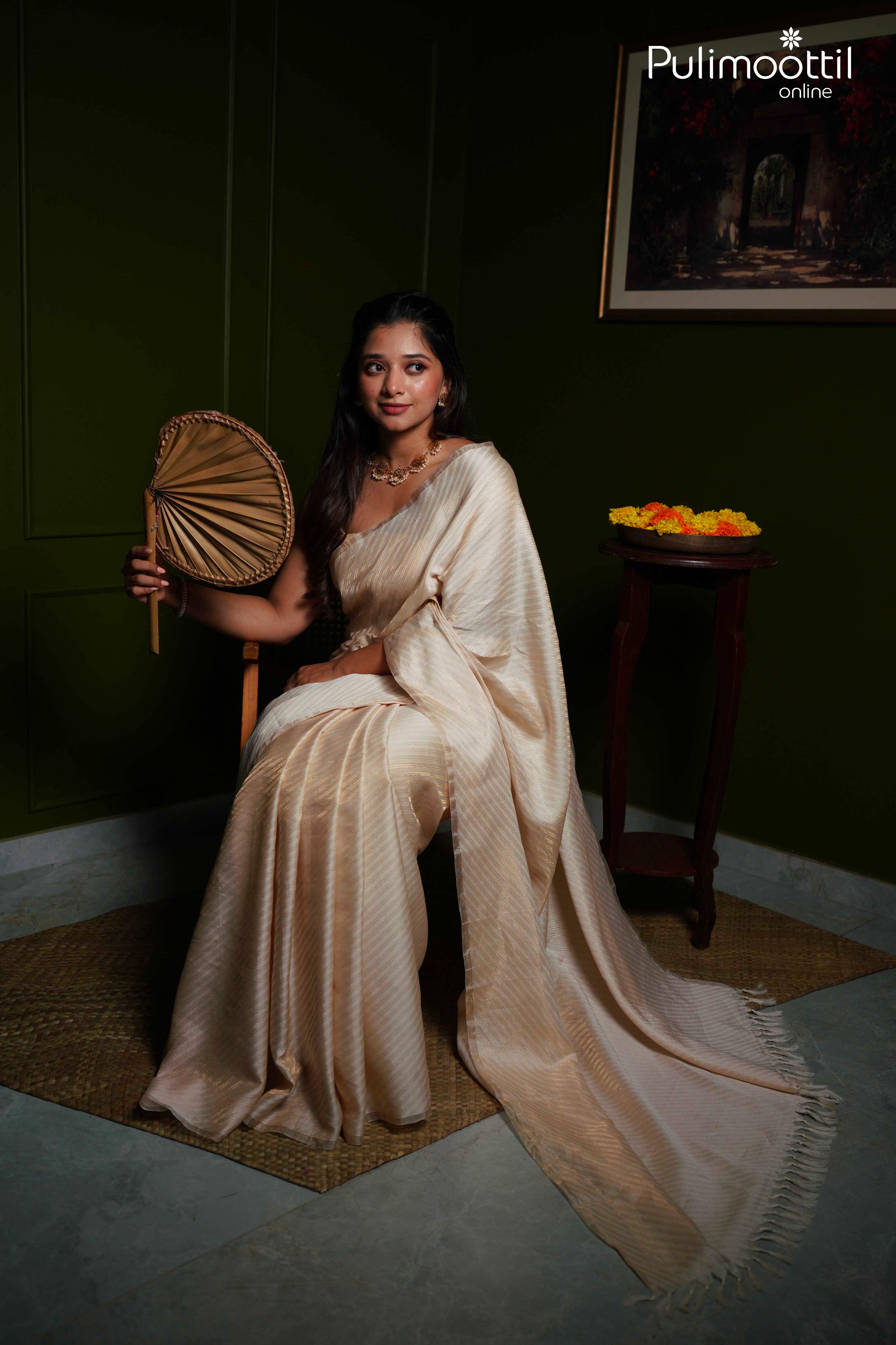 Traditional Indian woman posing in cream brocade saree, accessorized with gold necklace and earrings.