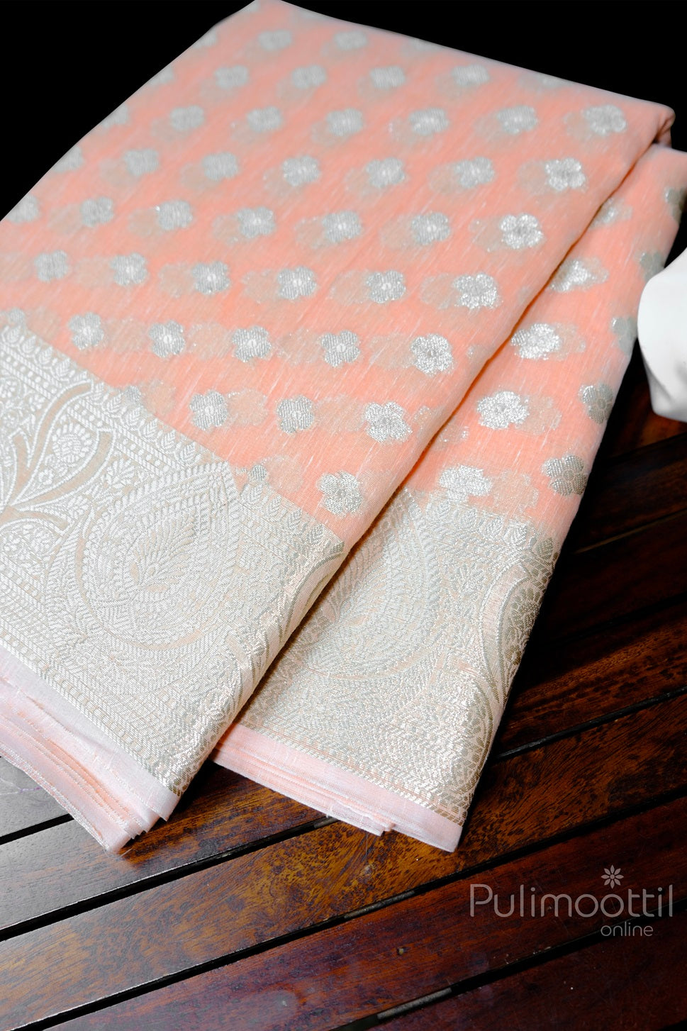 Creamy peach semi organza saree folded neatly with intricate silver zari floral motifs and elegant woven tissue border, placed on a wooden surface.