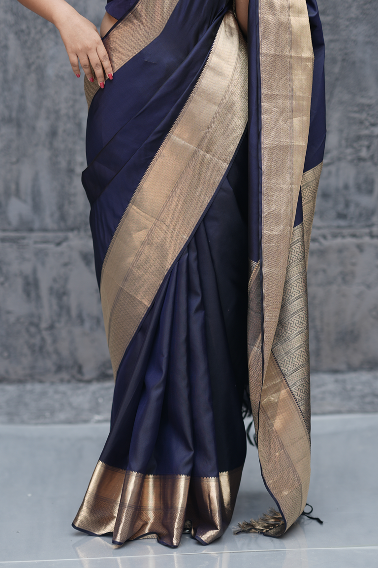 Close-up of hands adorned with a pearl ring holding the pallu of an ink blue Kanchipuram silk saree with golden zari border and intricate brocade work.