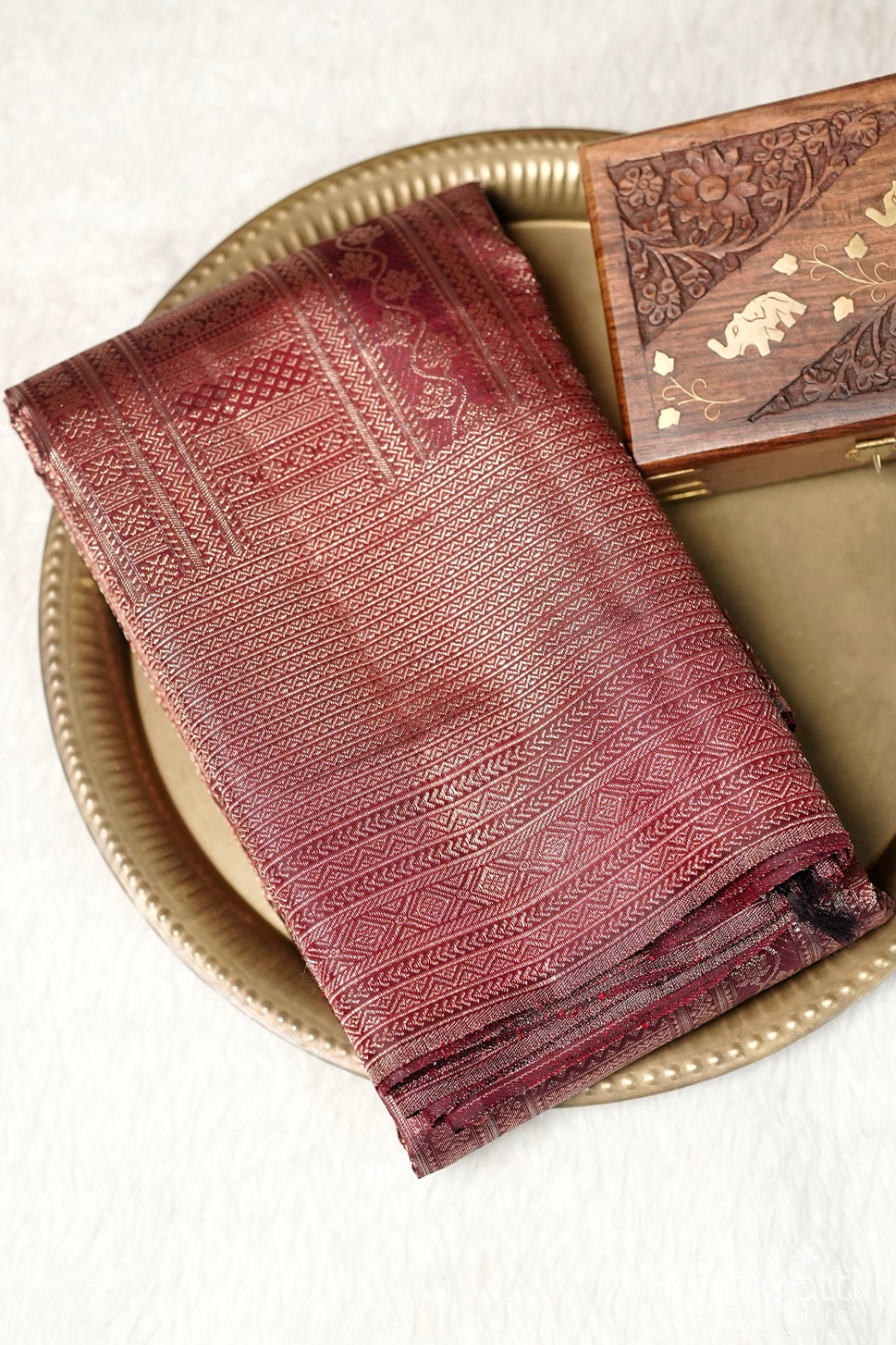 Dark maroon Kanchipuram brocade wedding saree with antique zari stripes and folded designer border placed on a golden plate with wooden box