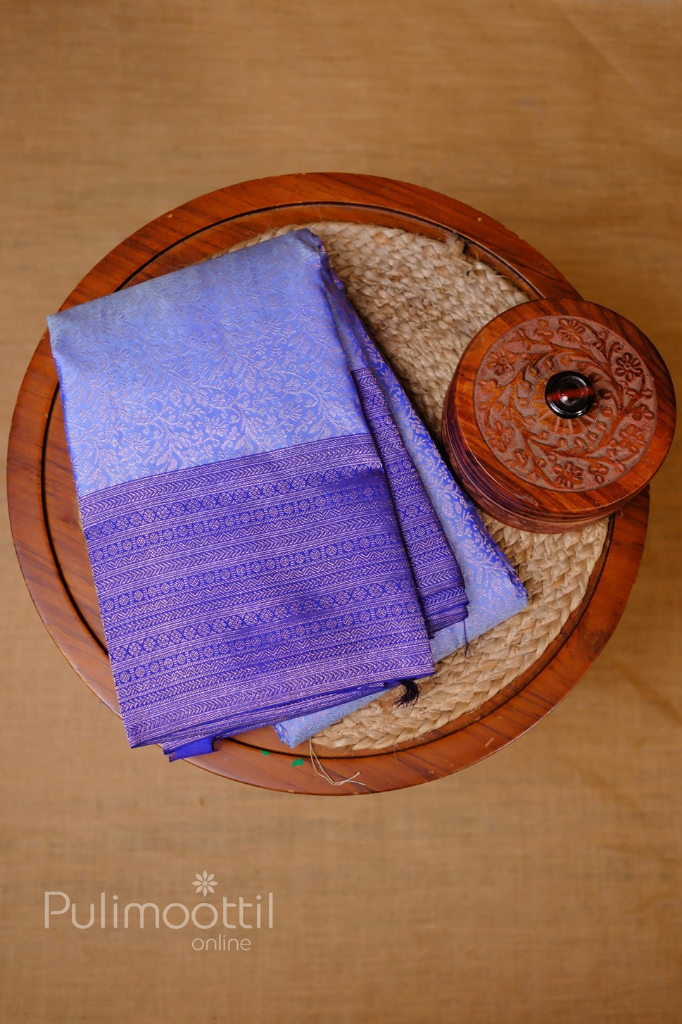 Folded dark purple Kanchipuram silk wedding saree with gold zari brocade border, styled on woven tray.
