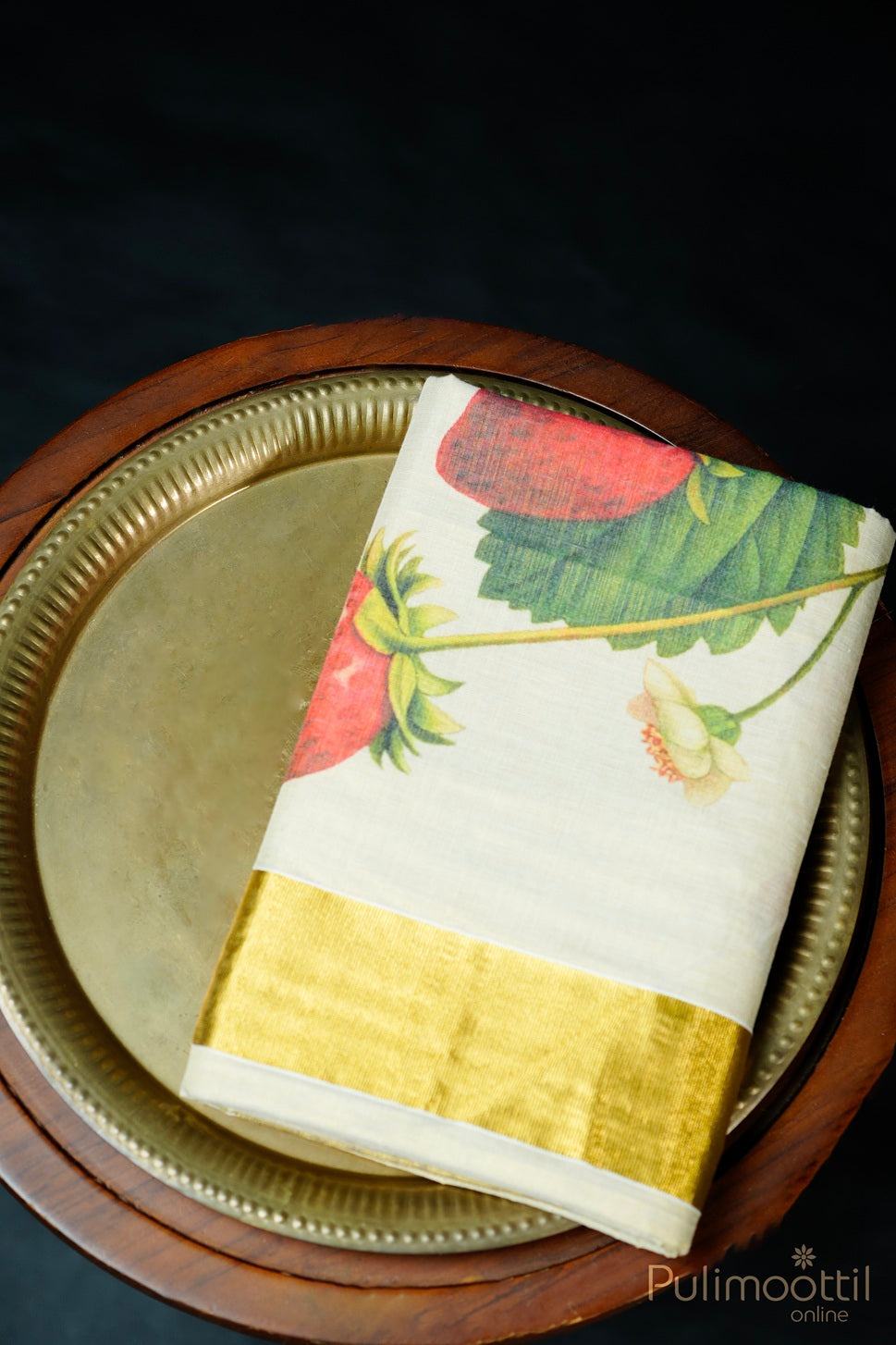 Folded golden tissue kasavu blouse piece with strawberry-pink fruit print near border, placed on a decorative brass plate.
