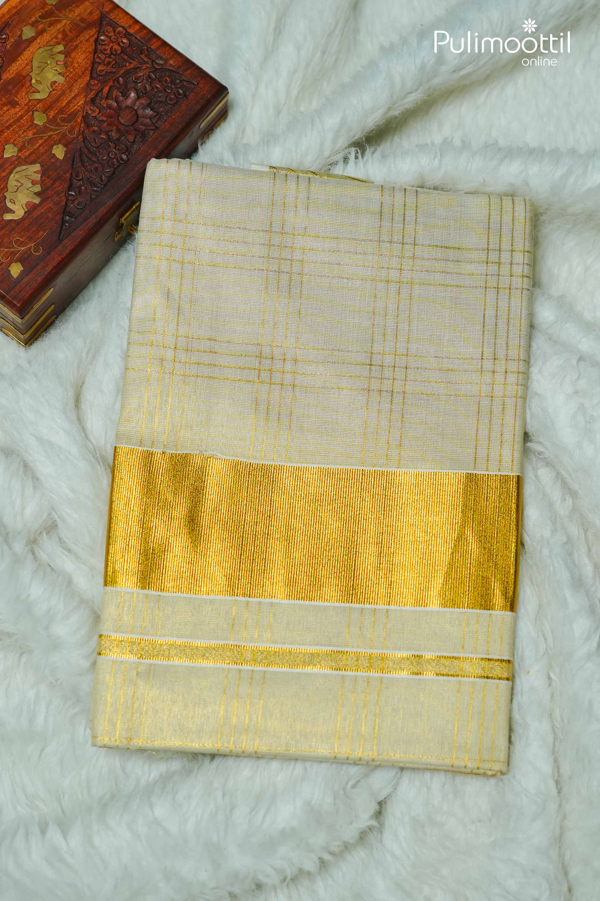 Folded golden zari check design tissue saree with broad golden kasavu border, placed on a soft white fabric background beside a carved wooden box – traditional Kerala set saree.