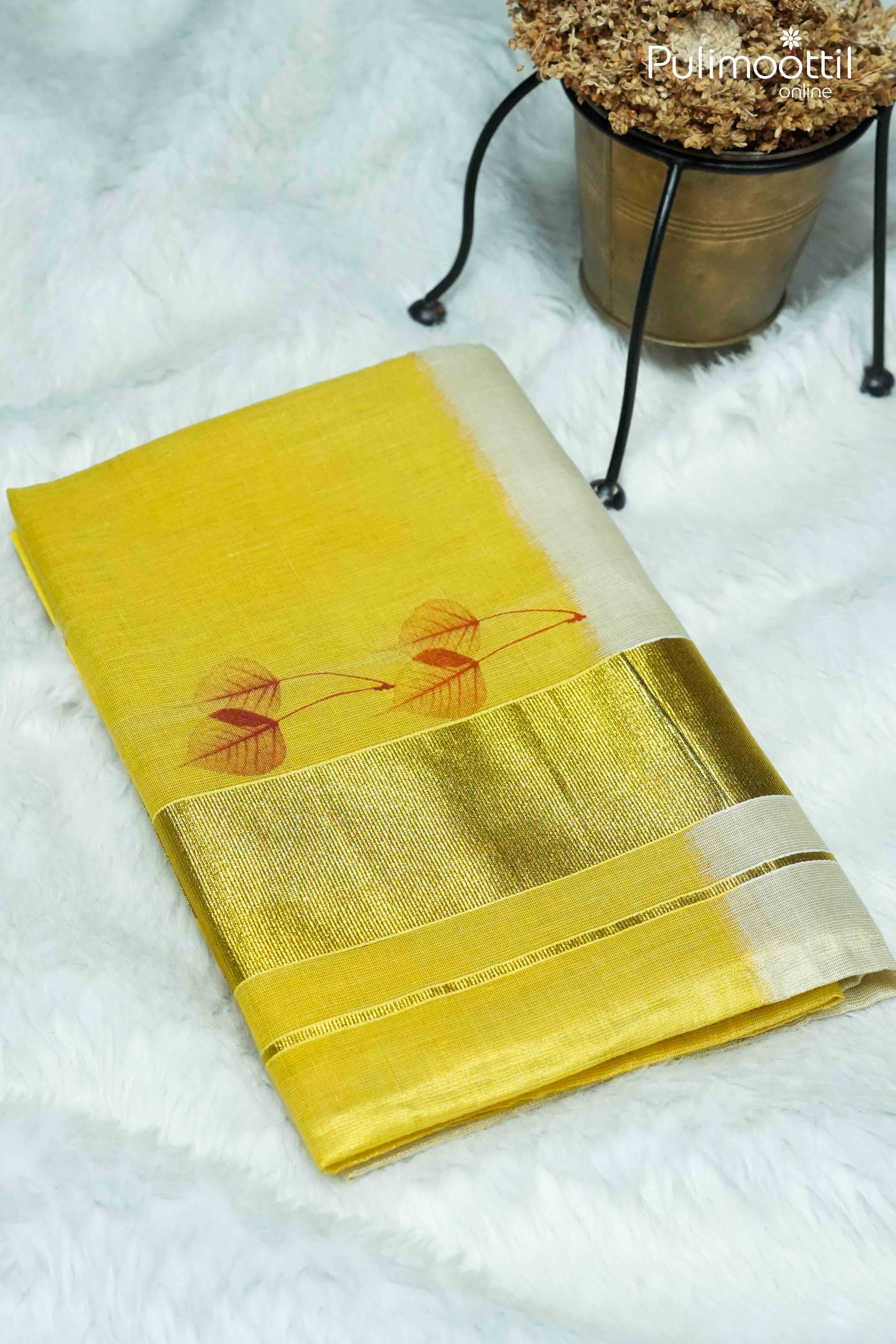 Folded Kerala golden tissue saree with metallic golden border and red leaf motifs, placed on white fur background.