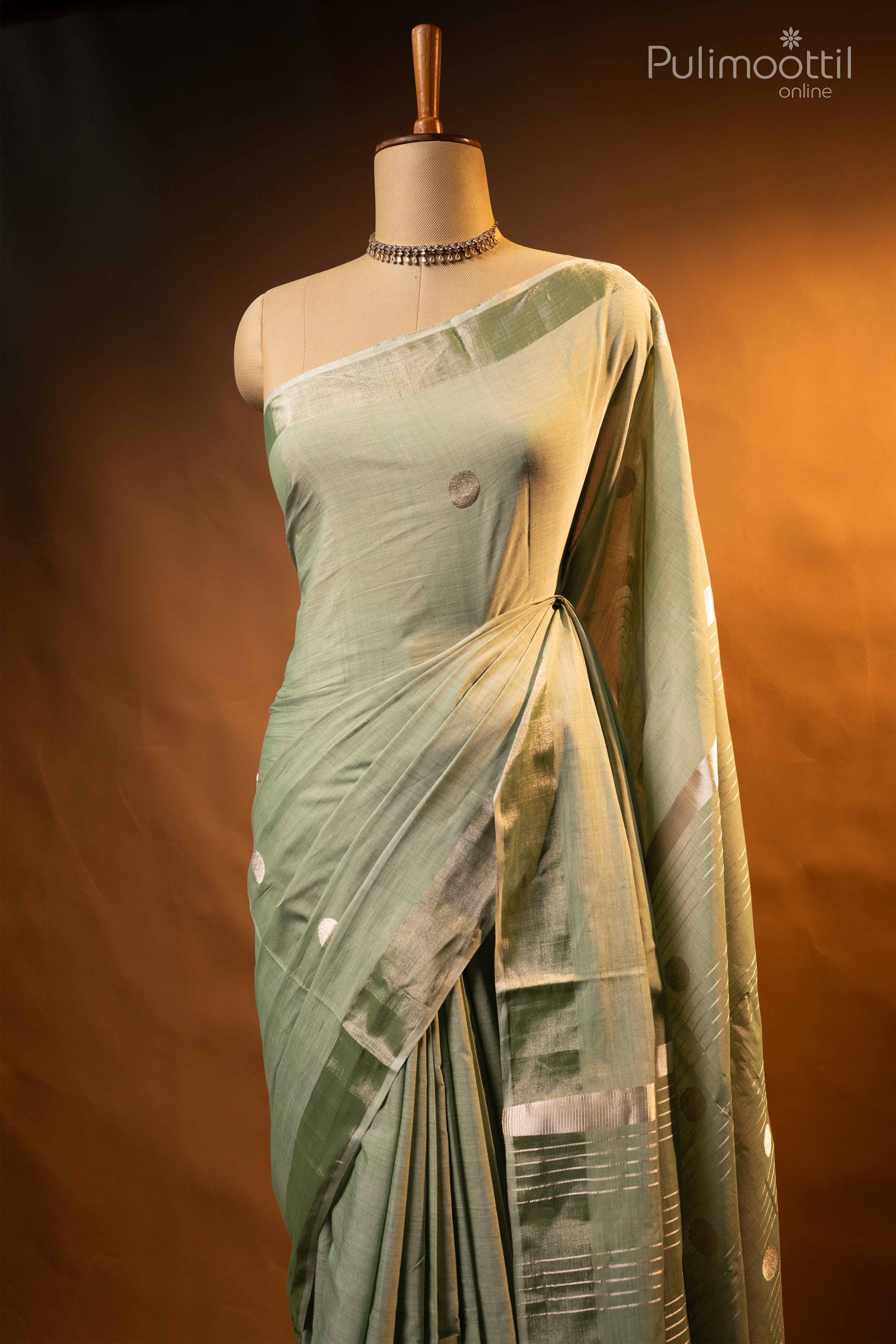 Light pista green handloom cotton saree draped on mannequin, adorned with silver zari buttas and tissue border, paired with elegant lighting backdrop.