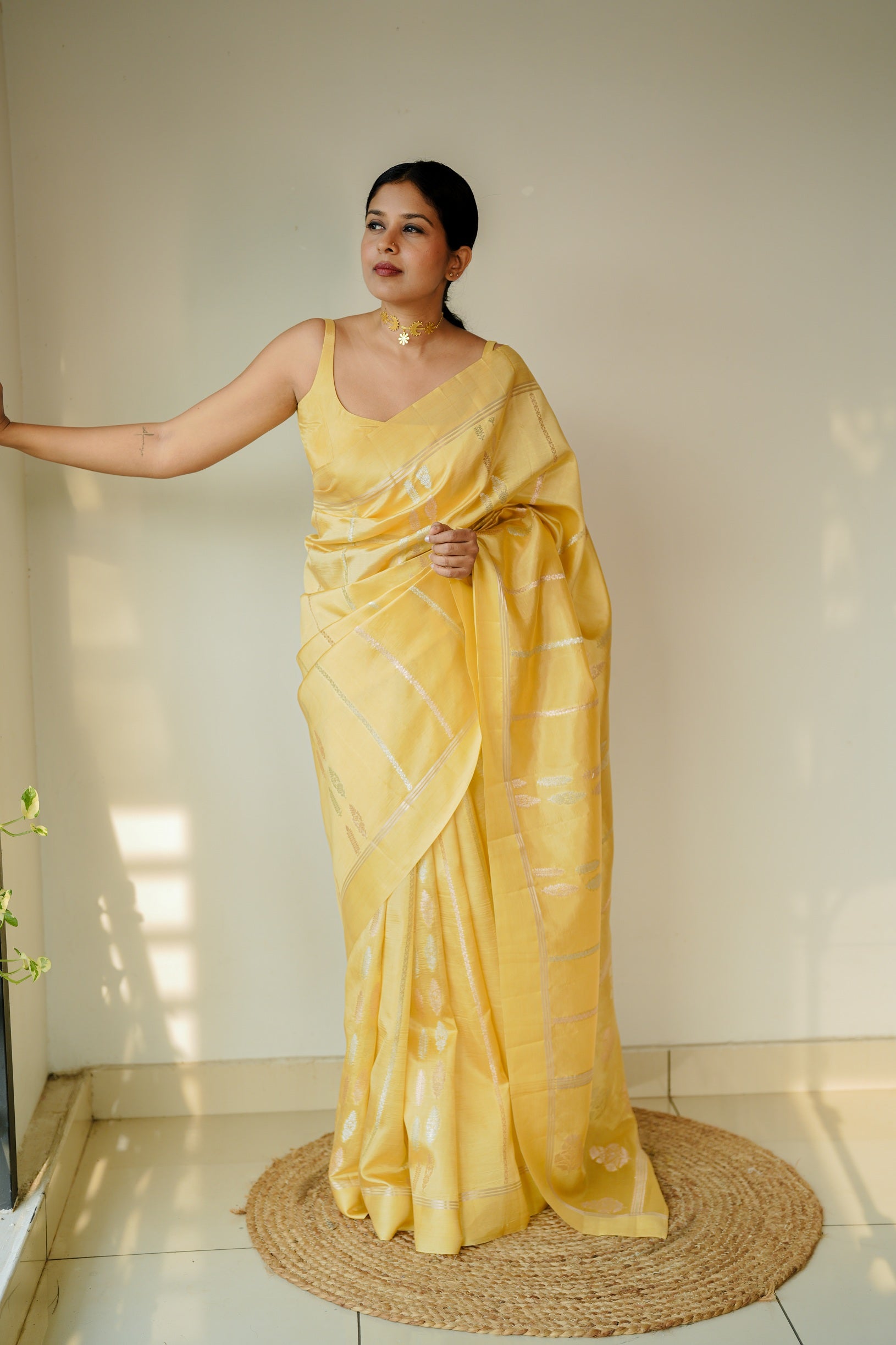 Full-length image of a woman standing in a yellow mango silk saree, styled with silver zari lines, looking to the side.
