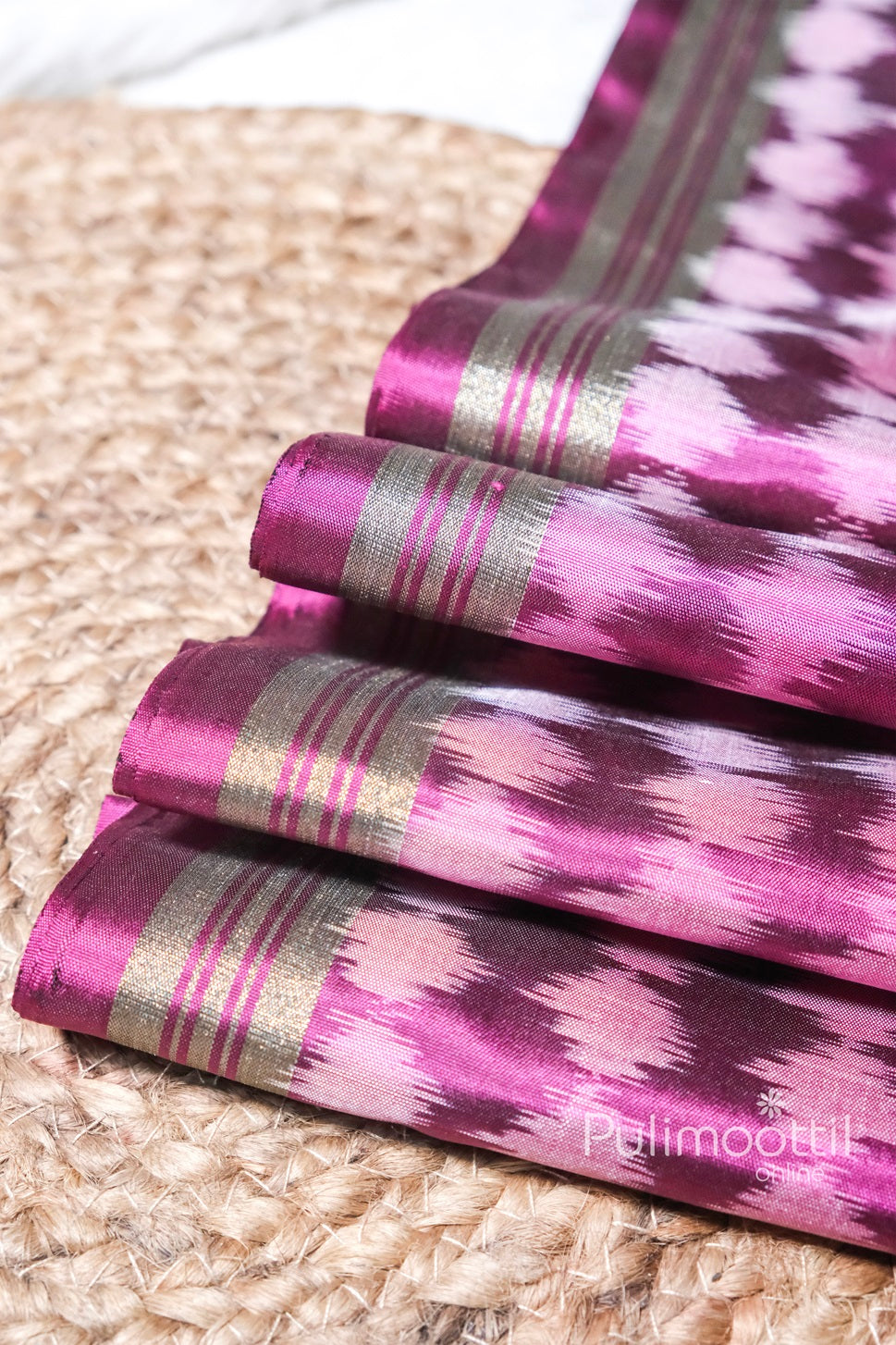 Folded view of magenta pure soft silk saree with pink and purple ikkat pattern, featuring antique golden zari border, displayed on a woven mat.
