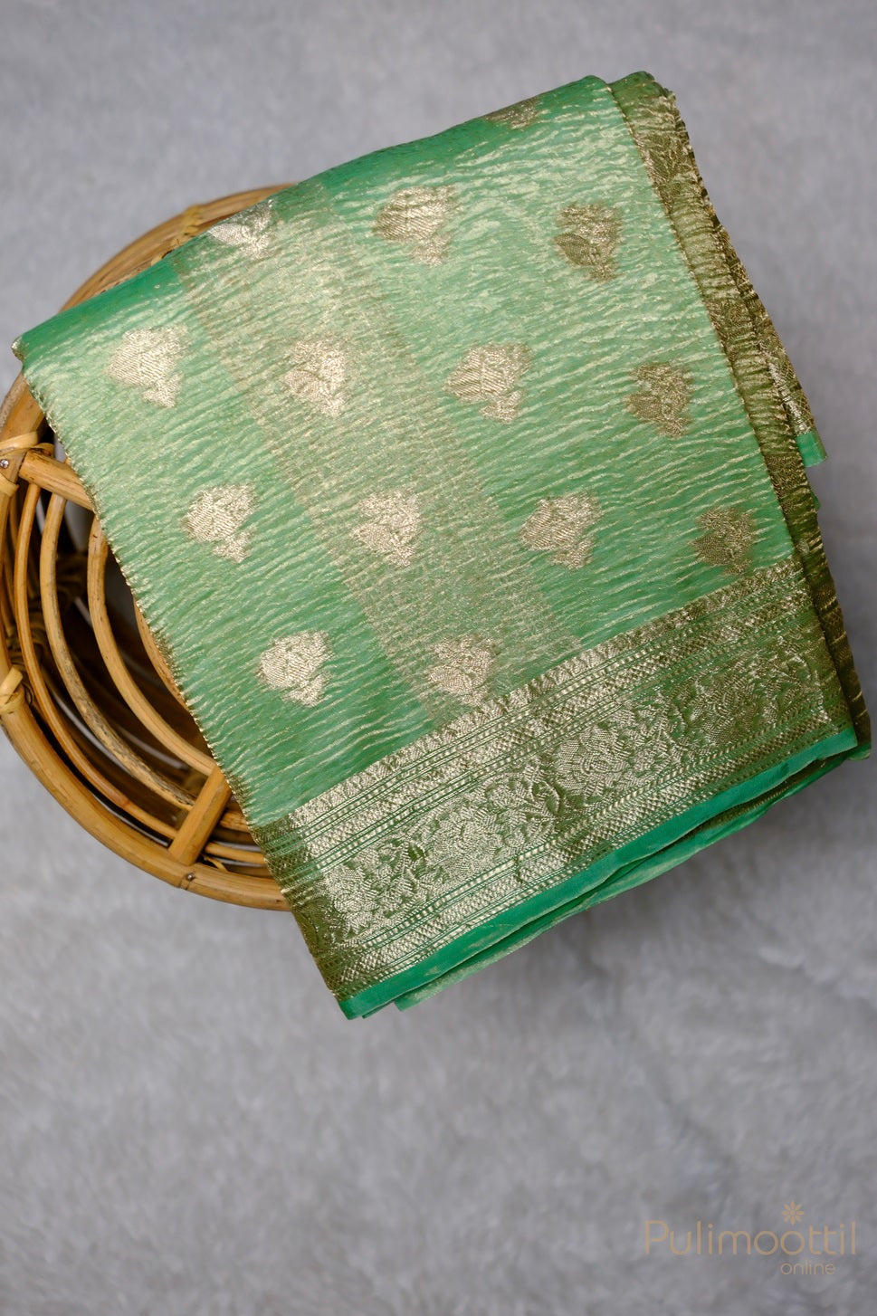 Mint green crushed Banarasi silk saree neatly folded in a basket, showcasing gold zari floral creeper designs and a delicate Banarasi border, highlighting the fabric’s luxurious sheen and intricate details.