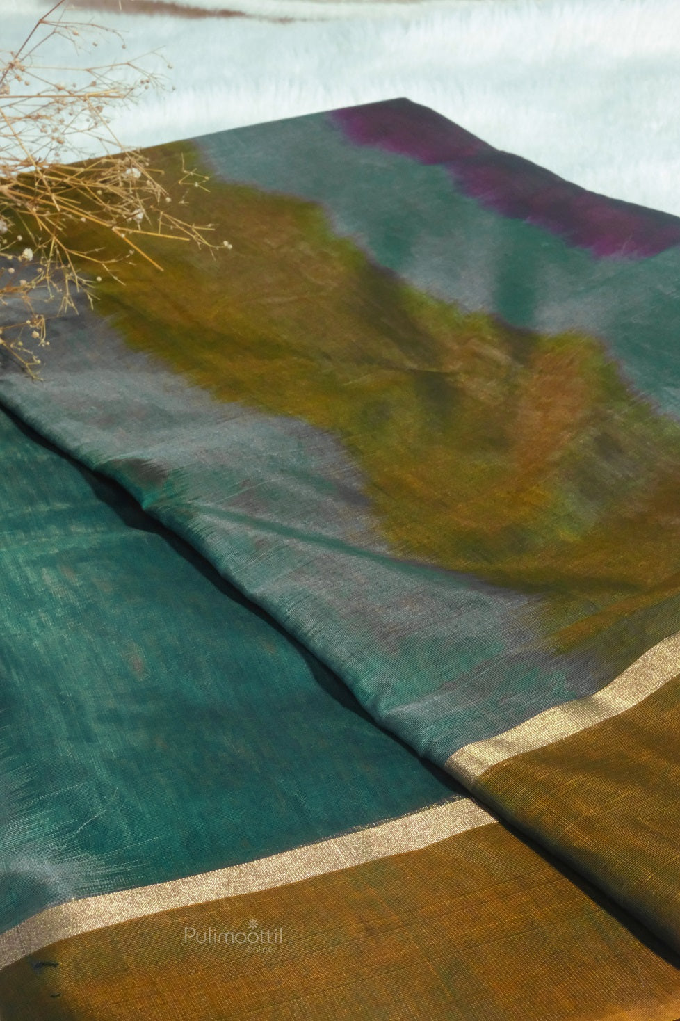 A close-up view of a multicolor cotton-silk saree with abstract dye patterns in green, mustard yellow, and purple tones. The fabric has a soft sheen and a minimal gold zari border.